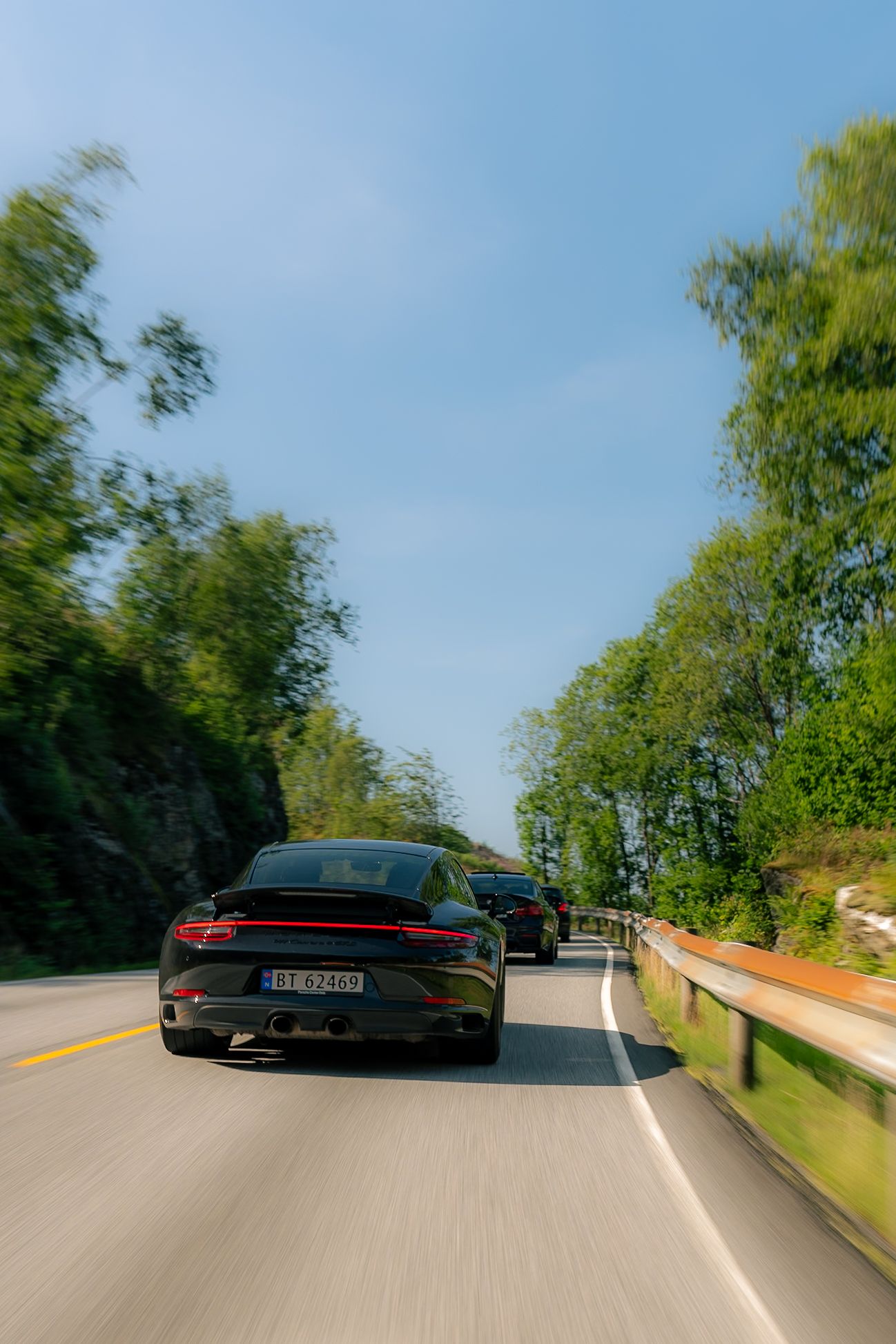 Black Porsche sports car driving along a winding scenic road lined with green trees and a guardrail under a clear blue sky.