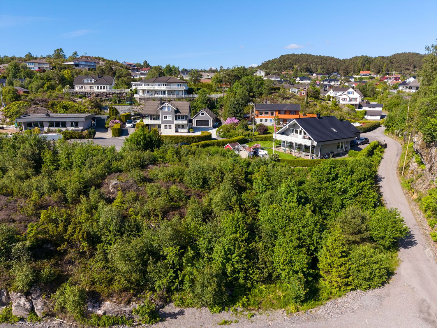 Aerial view of a hillside residential neighborhood with modern homes, lush green trees, and a winding gravel road under a clear blue sky.