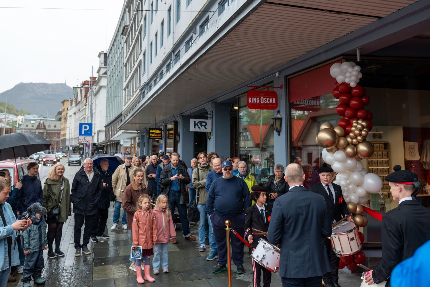 Crowd of people in coats and umbrellas gathers on a wet city street outside a decorated shop with red and gold balloons.