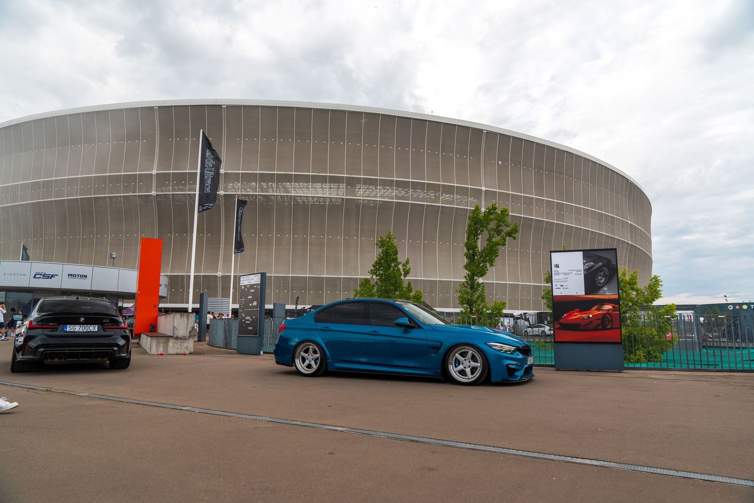 Blue BMW M car parked outside modern stadium building with curved architecture.