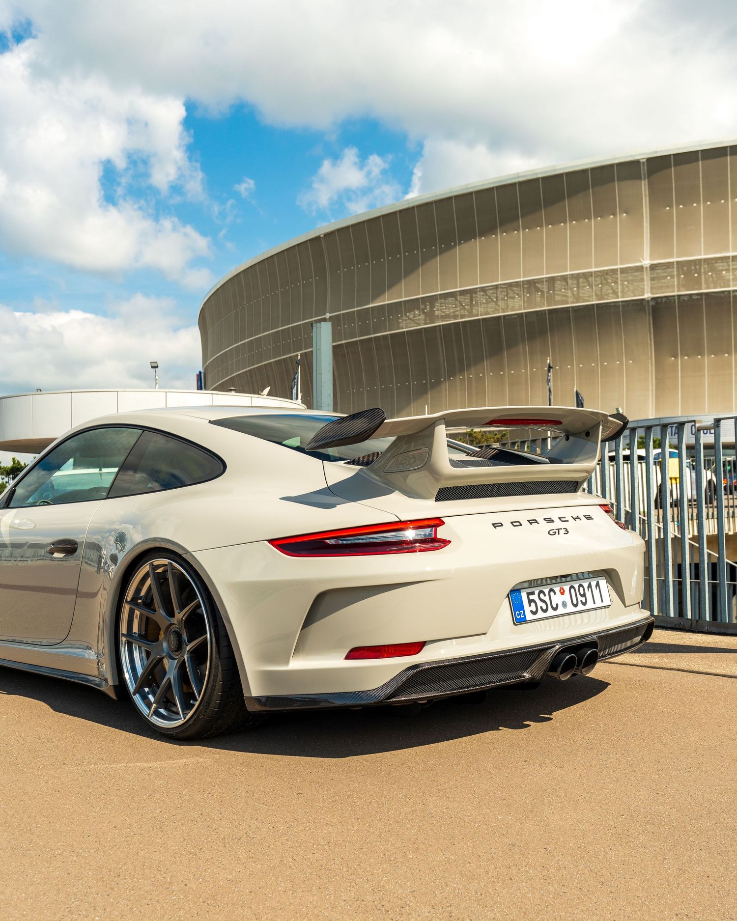 A white Porsche 911 GT3 parked in front of a modern stadium building with blue sky and clouds.