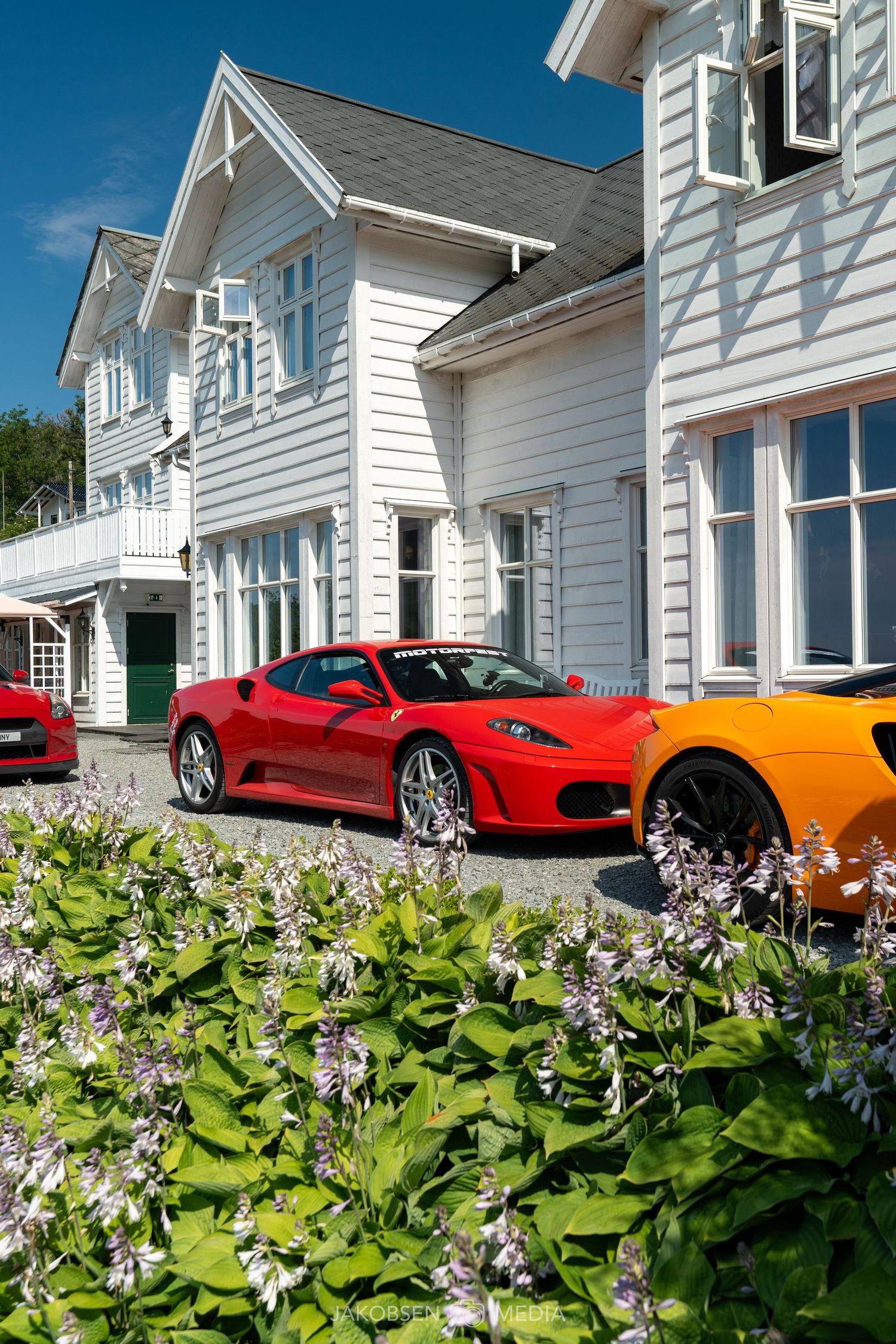 Luxury sports cars including red Ferrari models lined up in front of white colonial-style houses with flowering bushes.
