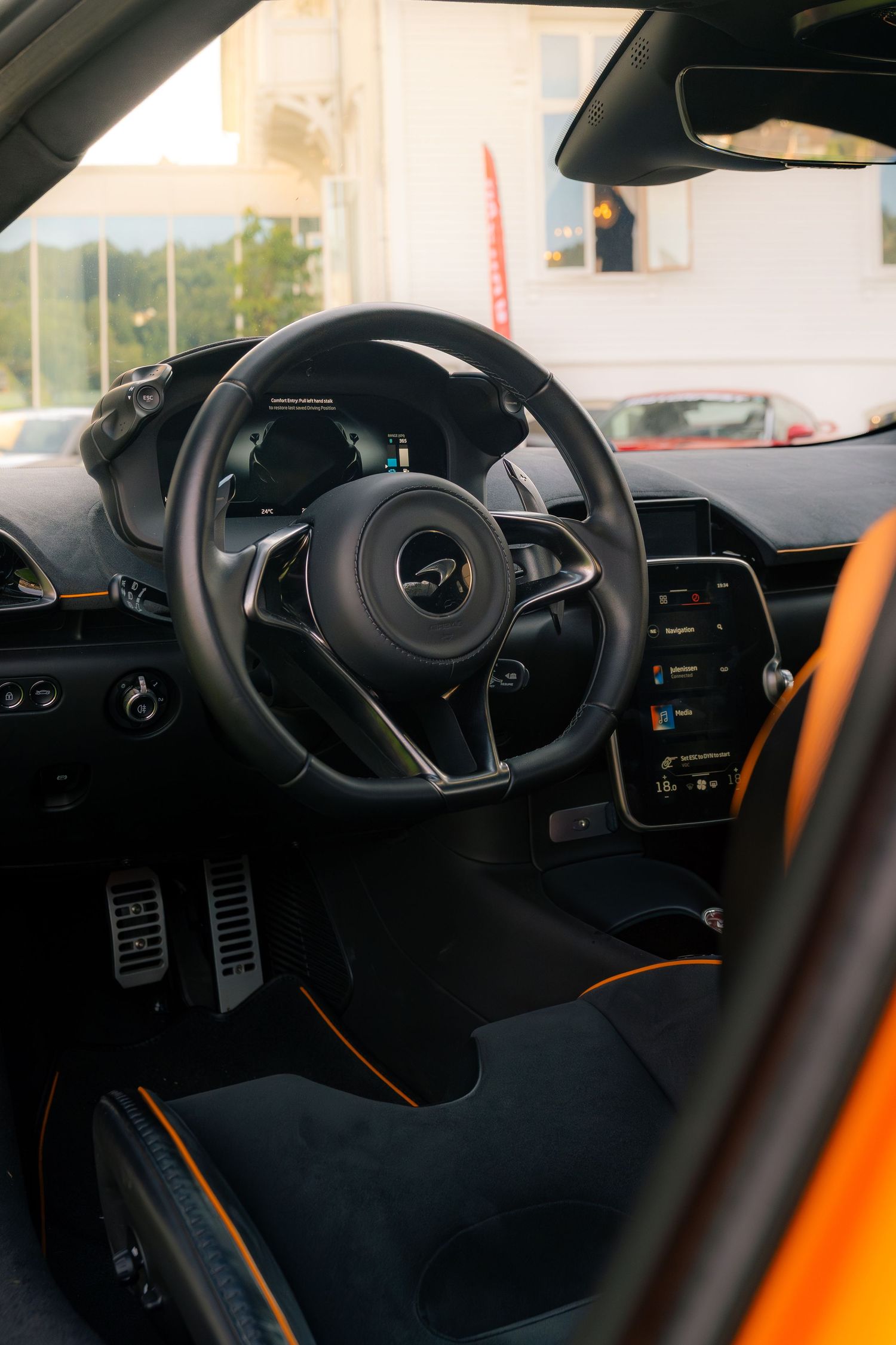 McLaren supercar interior featuring black leather steering wheel, orange accents, digital touchscreen, and aluminum pedals.