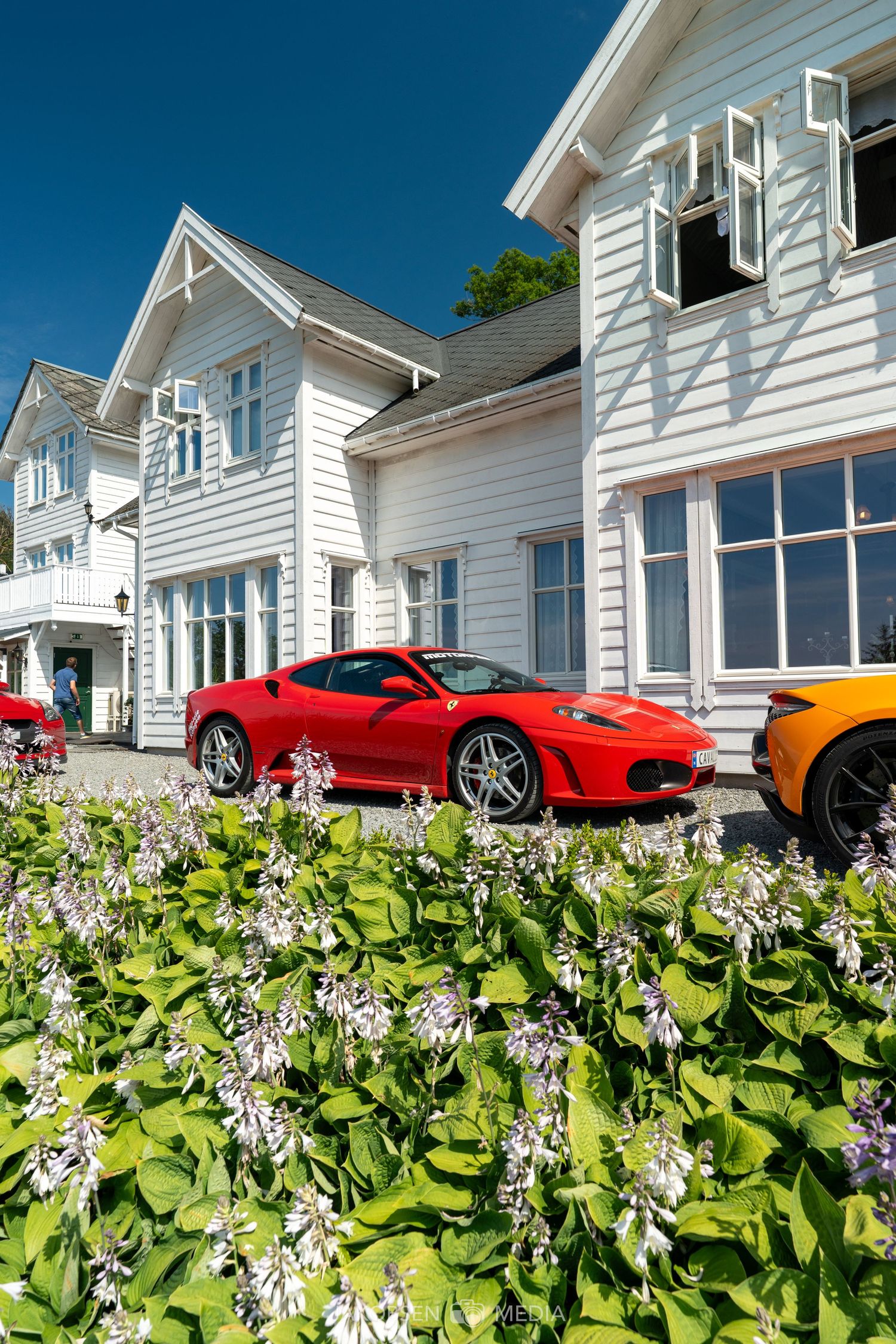 Luxury sports cars lined up in front of white cottages with blooming flowers in foreground.