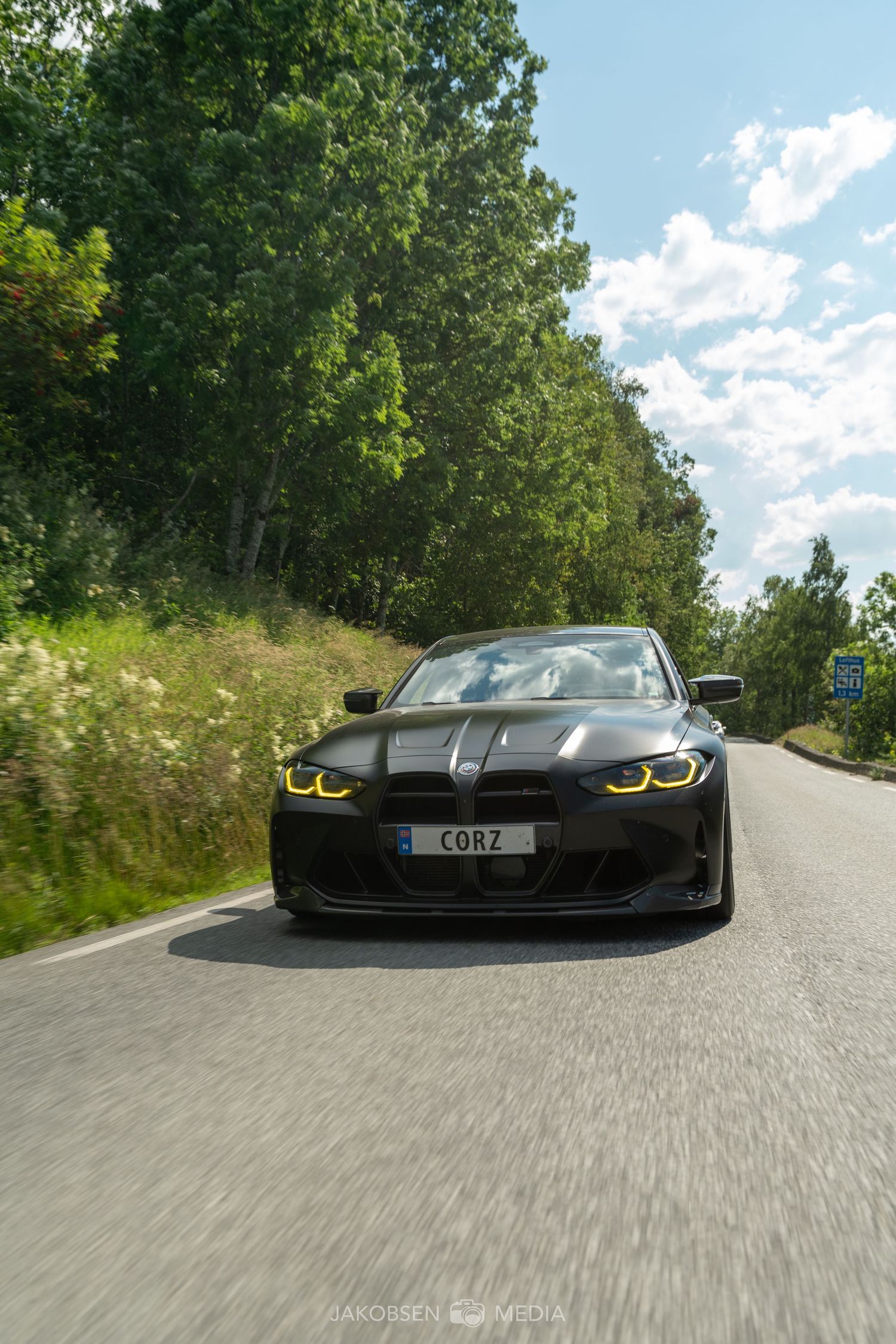 Front view of a sleek black BMW M4 driving on a winding forest road on a sunny day.
