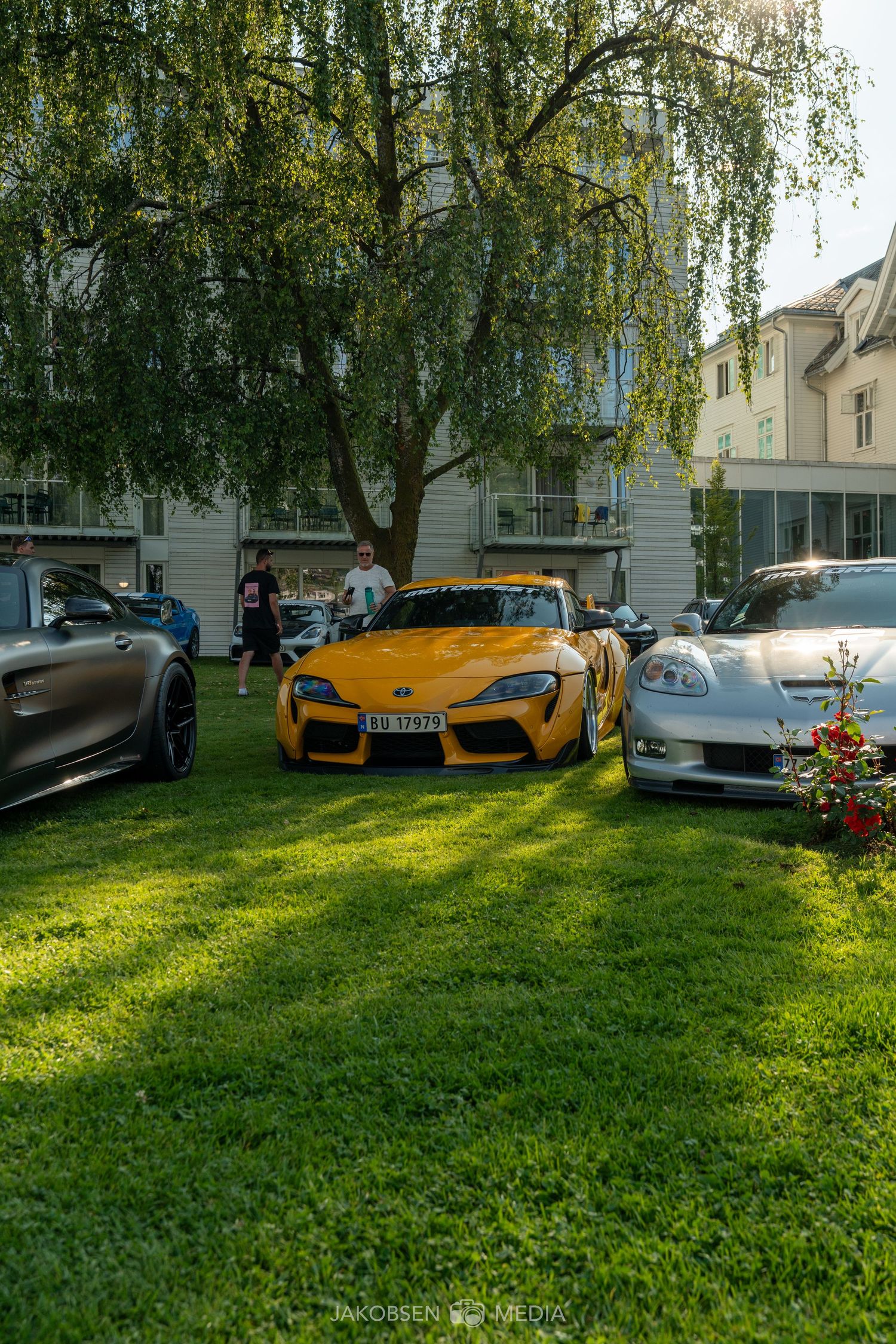 A bright yellow McLaren supercar parked on a grassy lawn next to trees and buildings in soft sunlight.