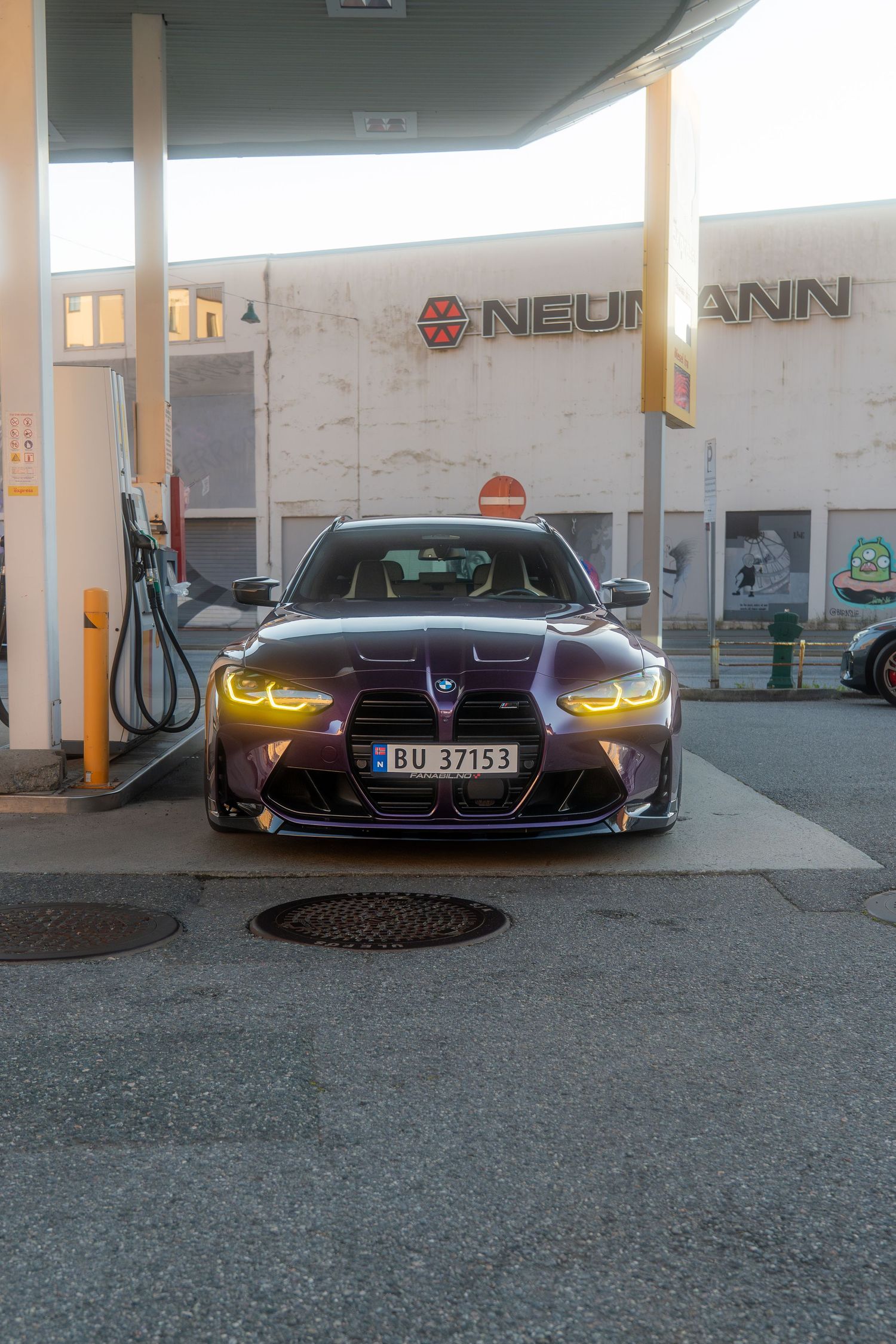 Purple BMW M4 with glowing yellow headlights parked under a gas station canopy at dusk.
