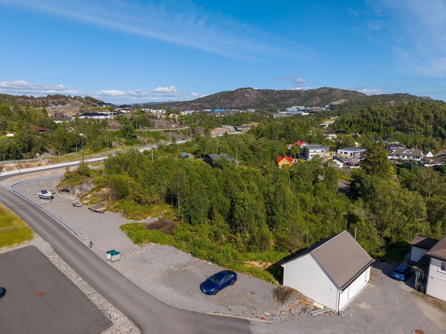 Hillside residential neighborhood with a white house, parked cars, and forested slopes beneath a blue sky with distant mountains.