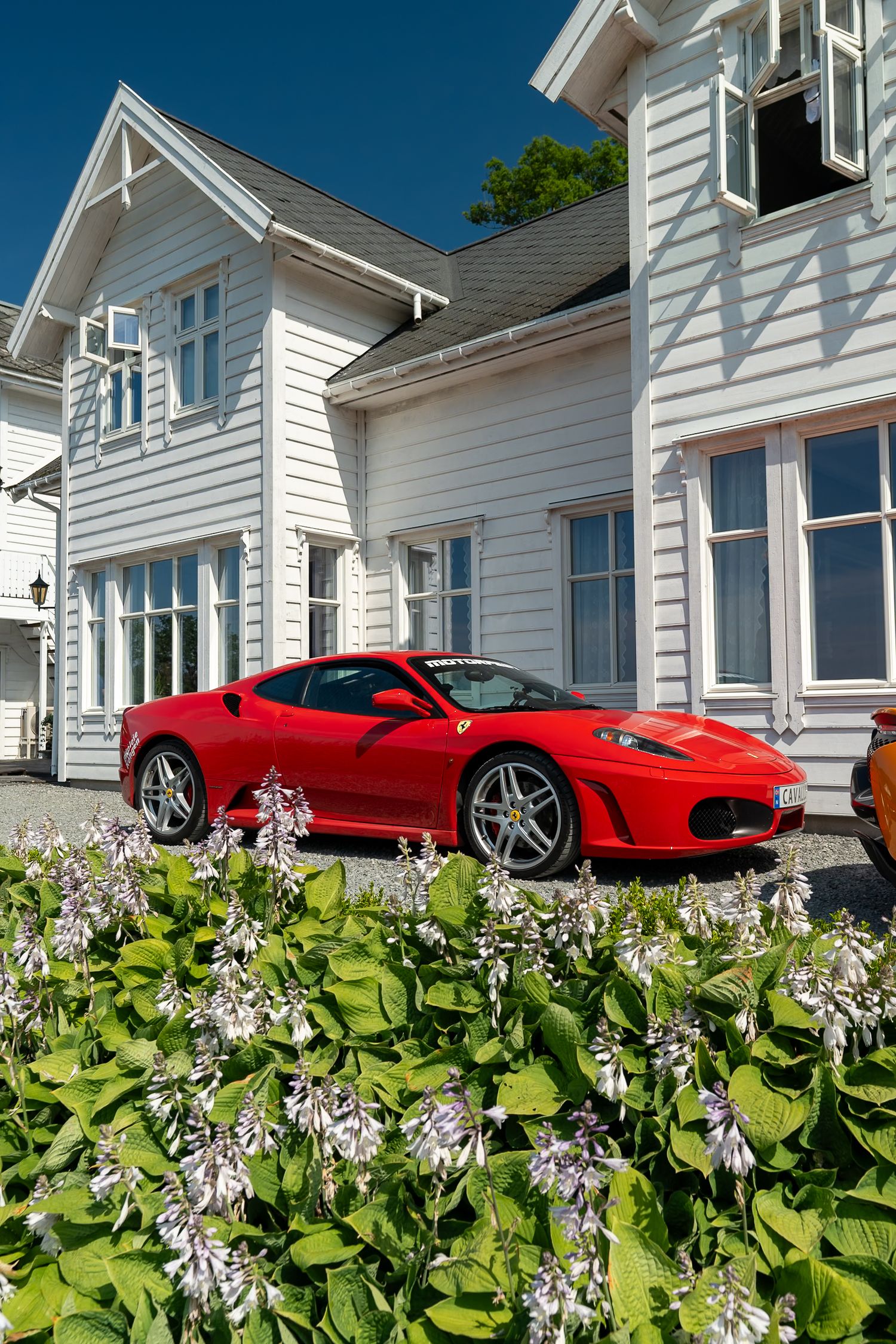 Red Ferrari sports car parked beside a white clapboard house with blooming hosta plants in the foreground on a sunny day.