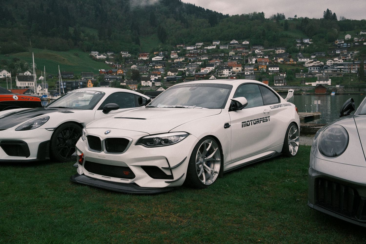 White BMW M2 sports car with a front splitter parked on grass at a car show, with a lakeside hillside village in the background.