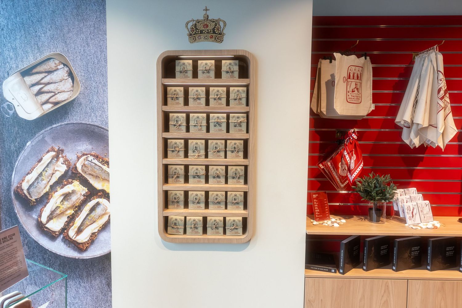 Wall-mounted wooden shelf with a crown emblem holding small boxed treats beside a sandwich photo and a red-paneled counter with tea towels.
