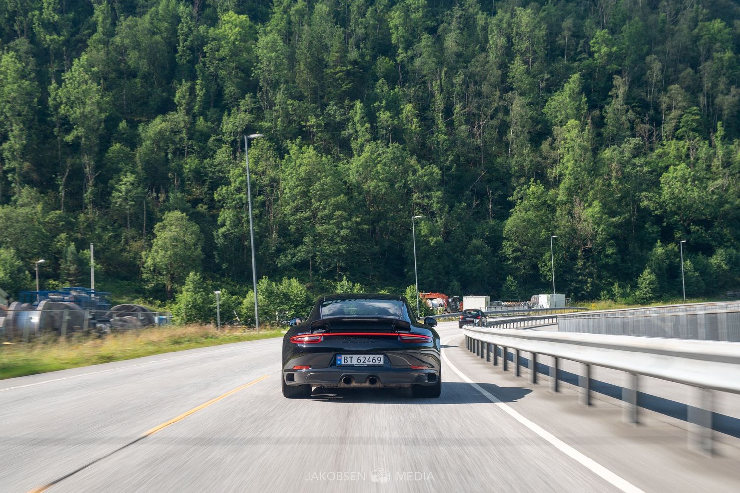 A winding mountain road curves through dense green forest with guard rails and clear pavement on a sunny day.