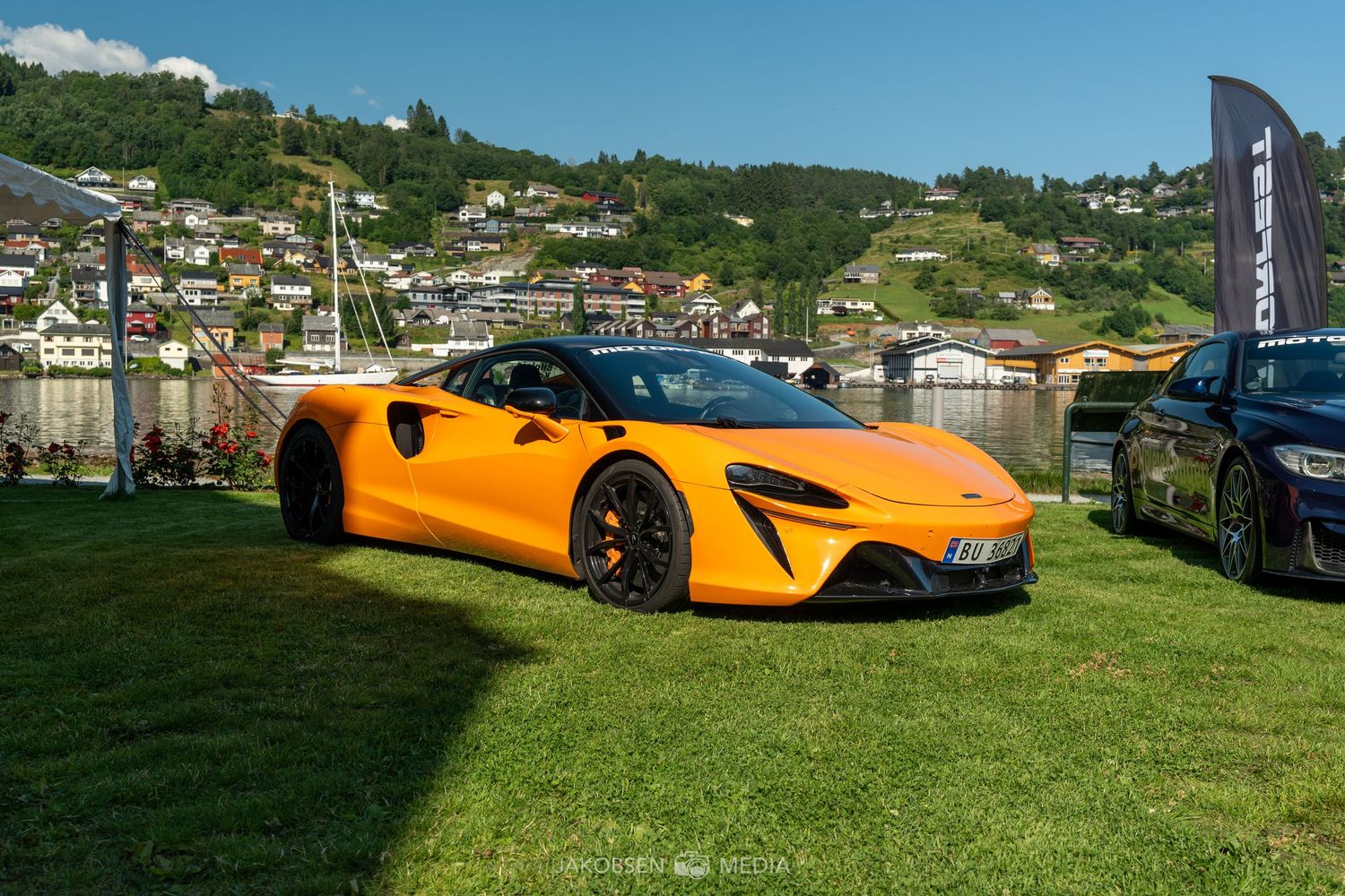 Orange McLaren supercars displayed on grass with mountain landscape in the background on a sunny day.