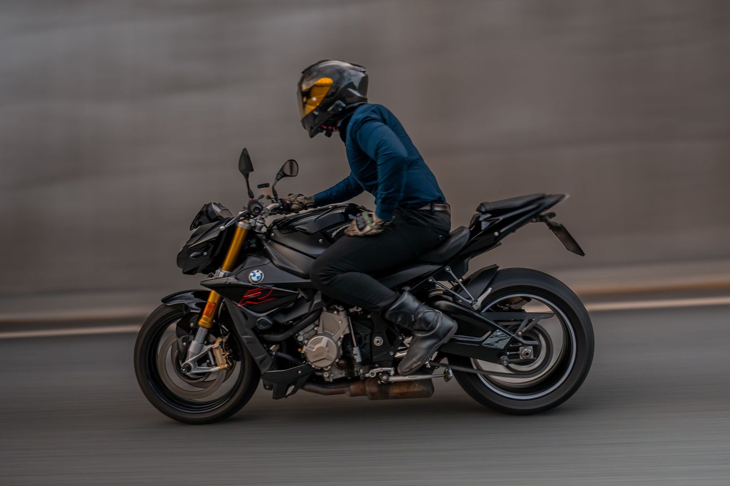 Motorcyclist in blue jacket rides a black BMW S1000R sportbike on a blurred motion road.
