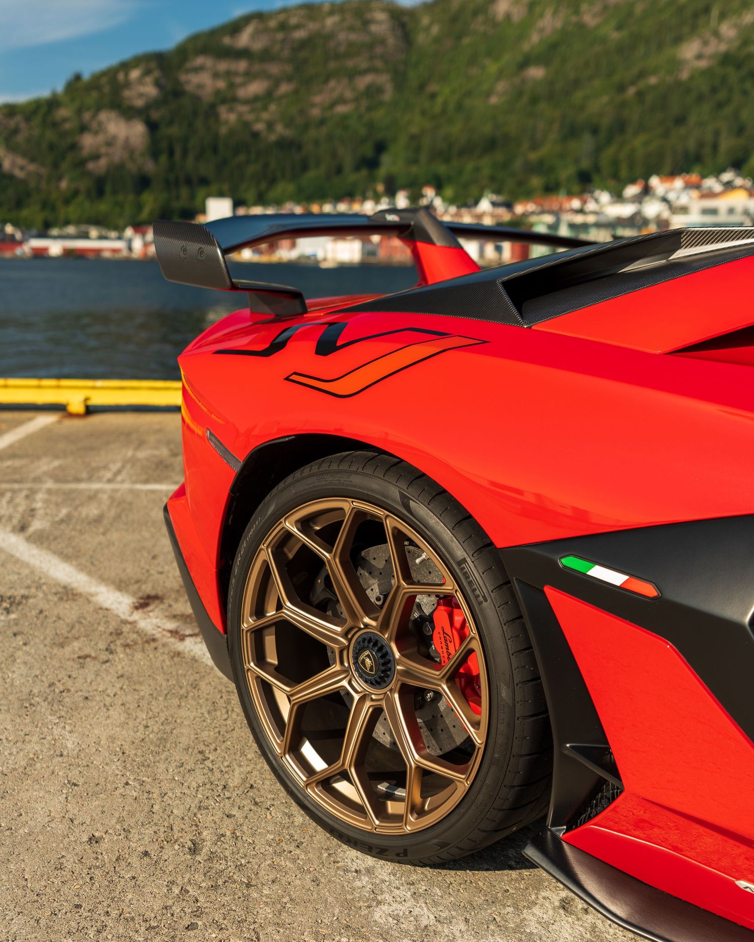 Red Lamborghini wheel and fender detail with bronze forged alloy wheel against coastal background.