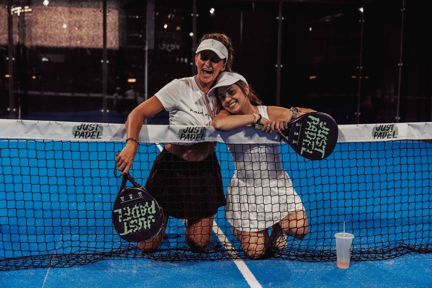 Two players resting against tennis net with padel rackets on blue court.