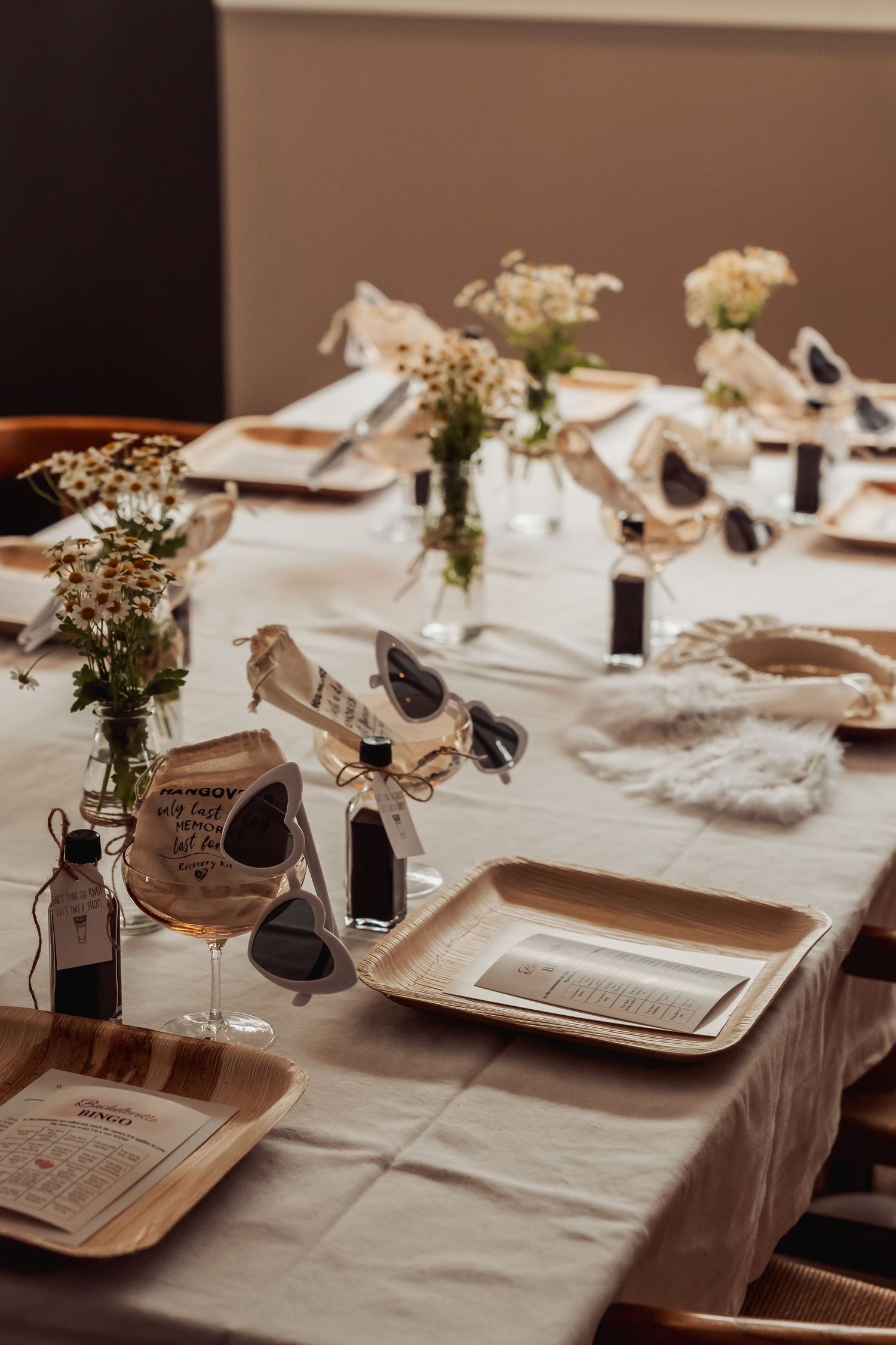 Rustic wedding table setting with sunglasses, white flowers in vases, and place settings on cream tablecloth.