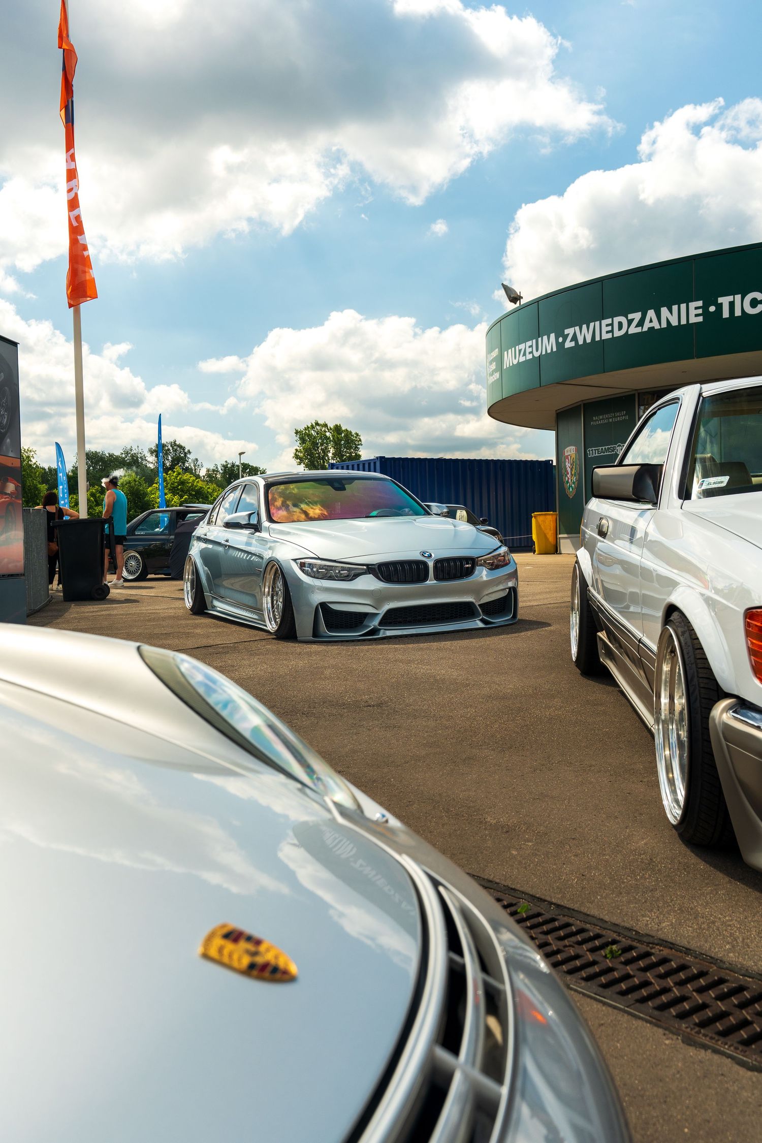 Lowered BMW M3 in silver parked at an outdoor event venue with other vehicles.