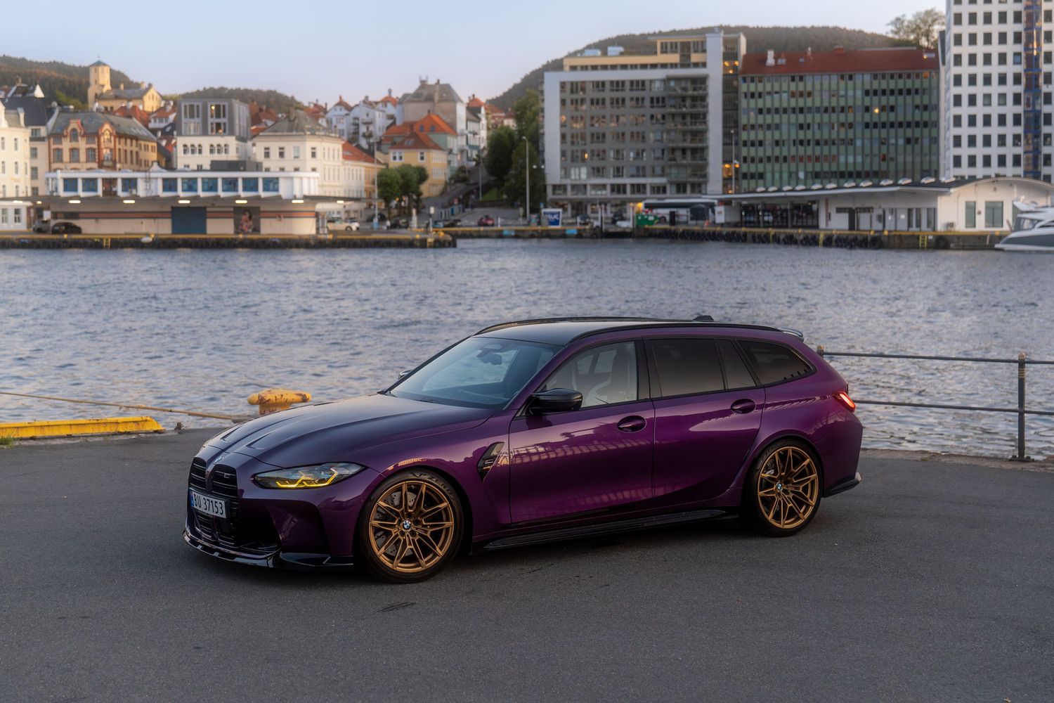 Purple BMW M3 Touring wagon parked along a waterfront with city buildings in the background.