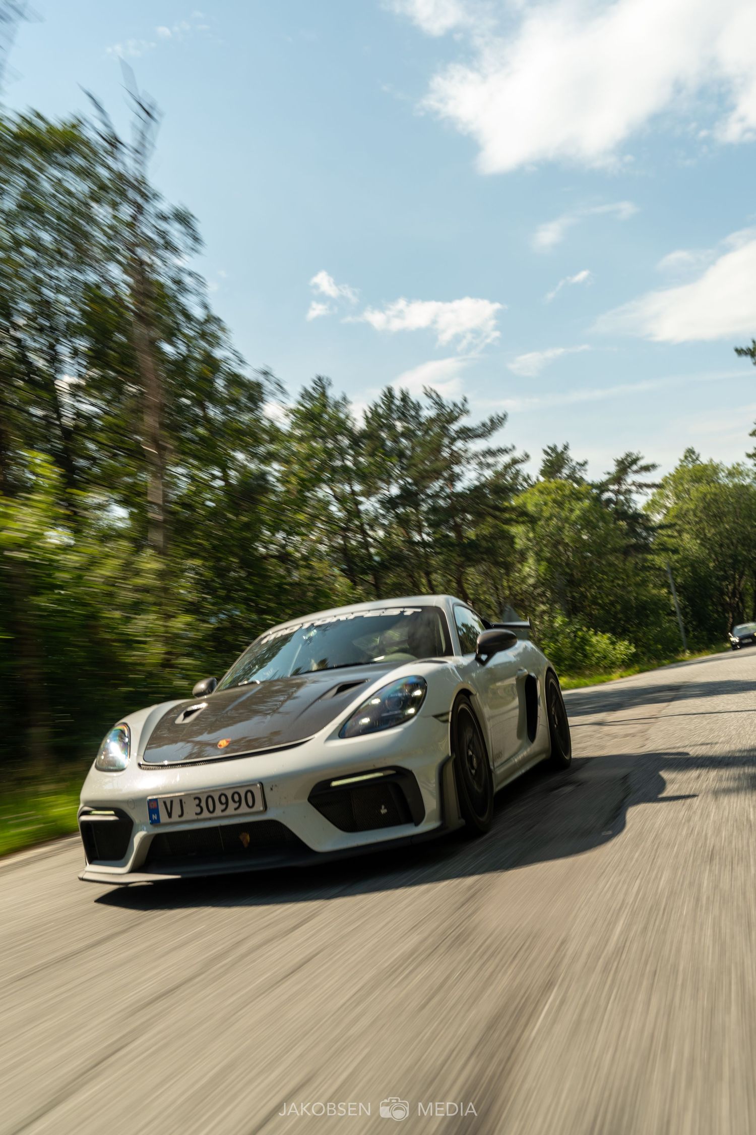 Silver Porsche Cayman GT4 driving on winding forest road with motion blur effect.