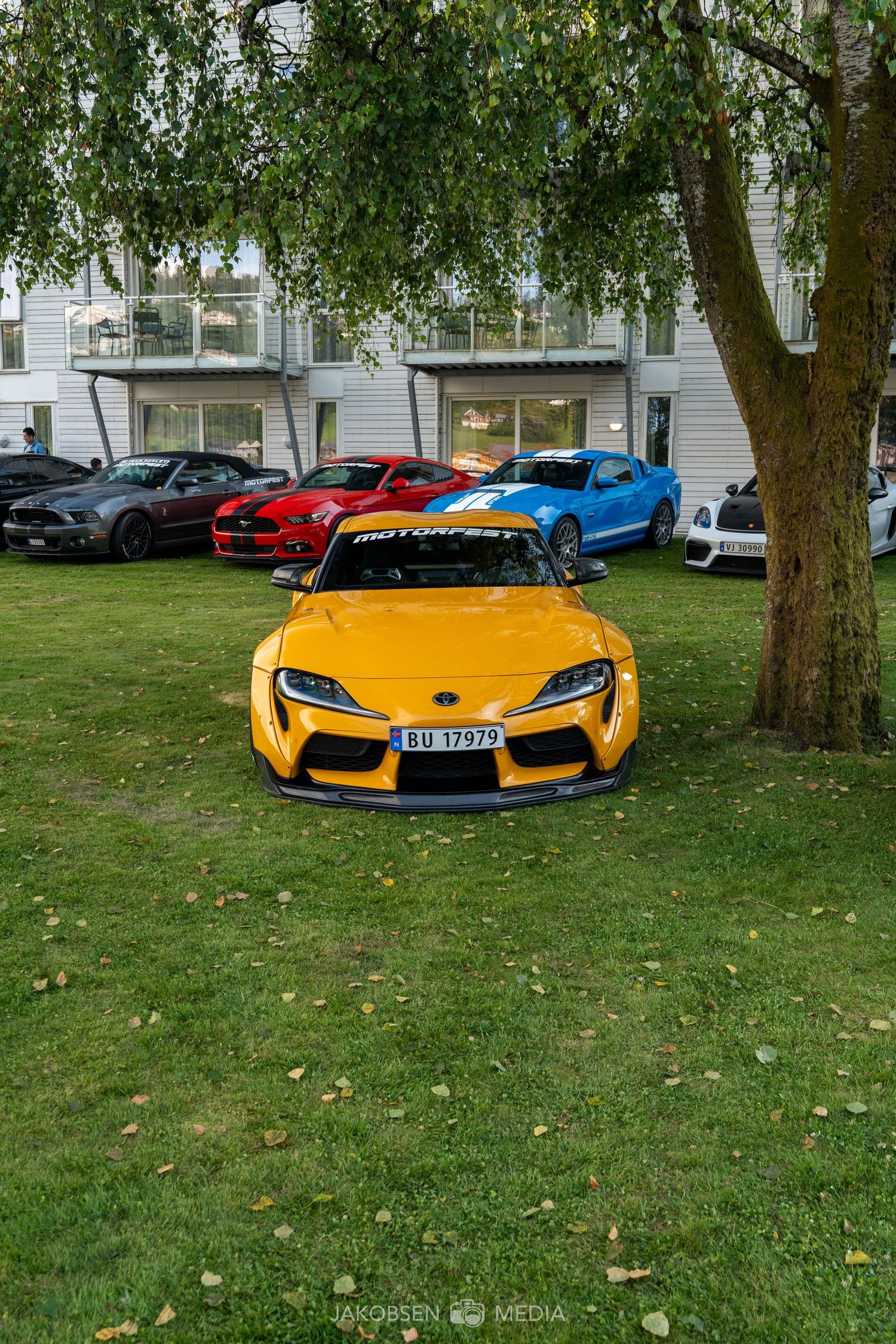 Bright yellow supercar parked on grass near trees and other luxury vehicles in a residential area.