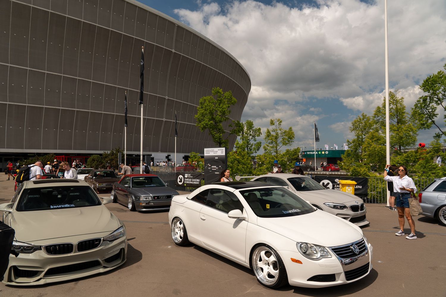 Luxury cars parked outside a large modern stadium building with curved architecture under cloudy skies.