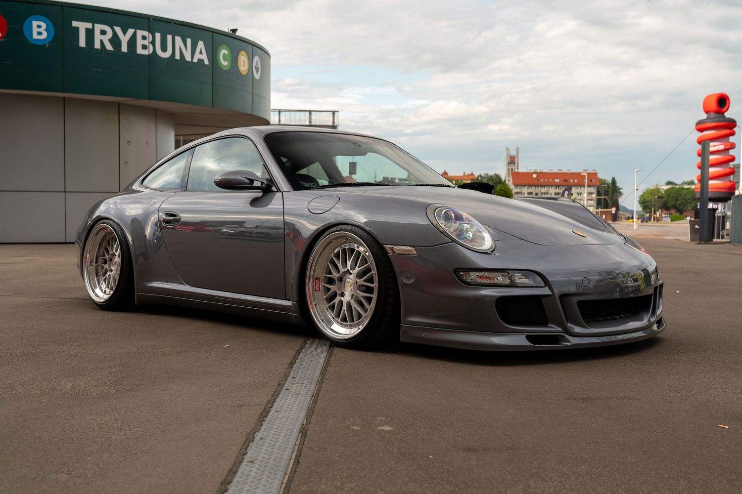 Modified grey Porsche 911 with lowered suspension and custom BBS wheels parked in front of Trybuna building.