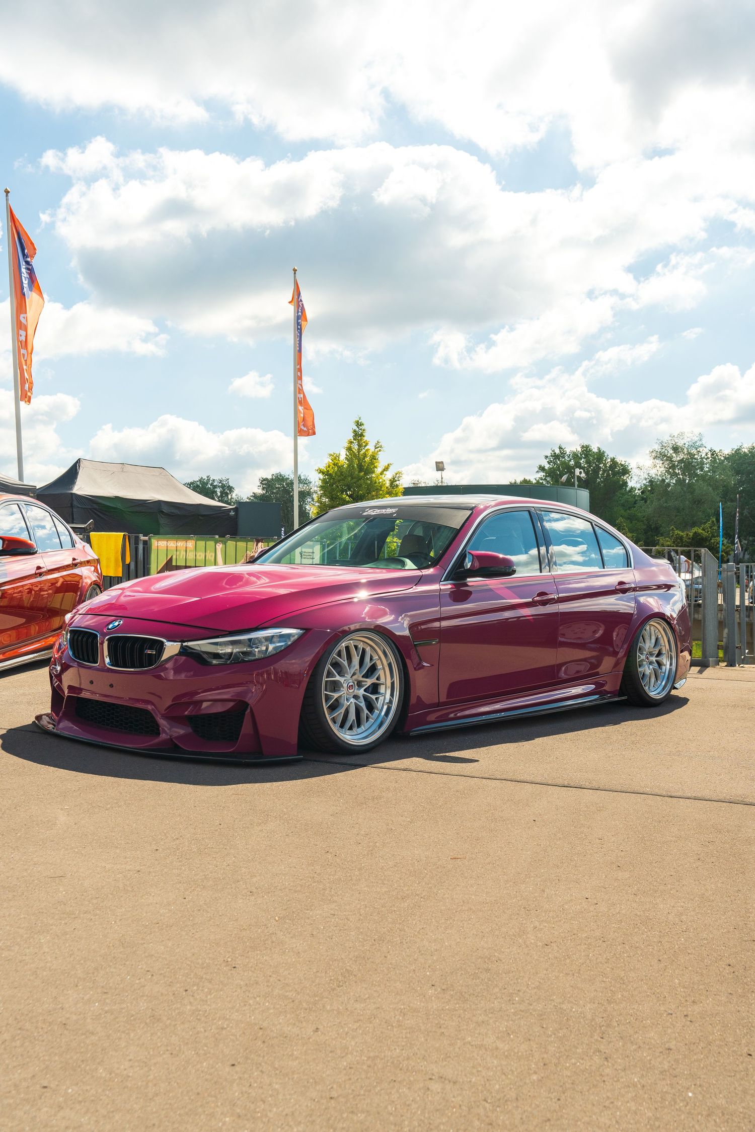 Customized burgundy BMW M3 sedan with lowered suspension and aftermarket wheels parked at a car show.