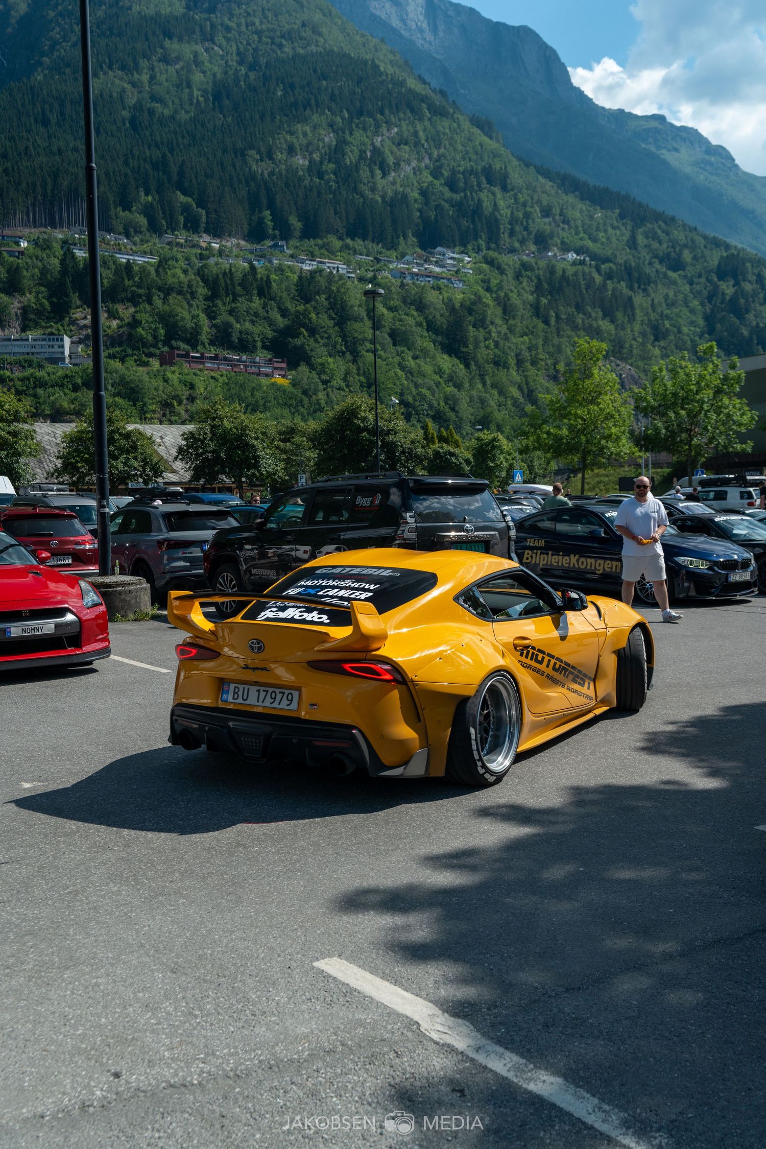Modified yellow sports car with wide body kit parked in mountain parking lot with scenic backdrop.