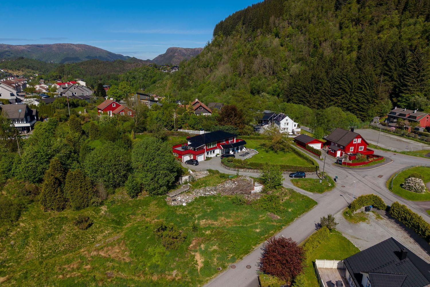 Aerial view of red and white houses nestled among green trees and lawns at the base of a forested mountain slope in a scenic village.