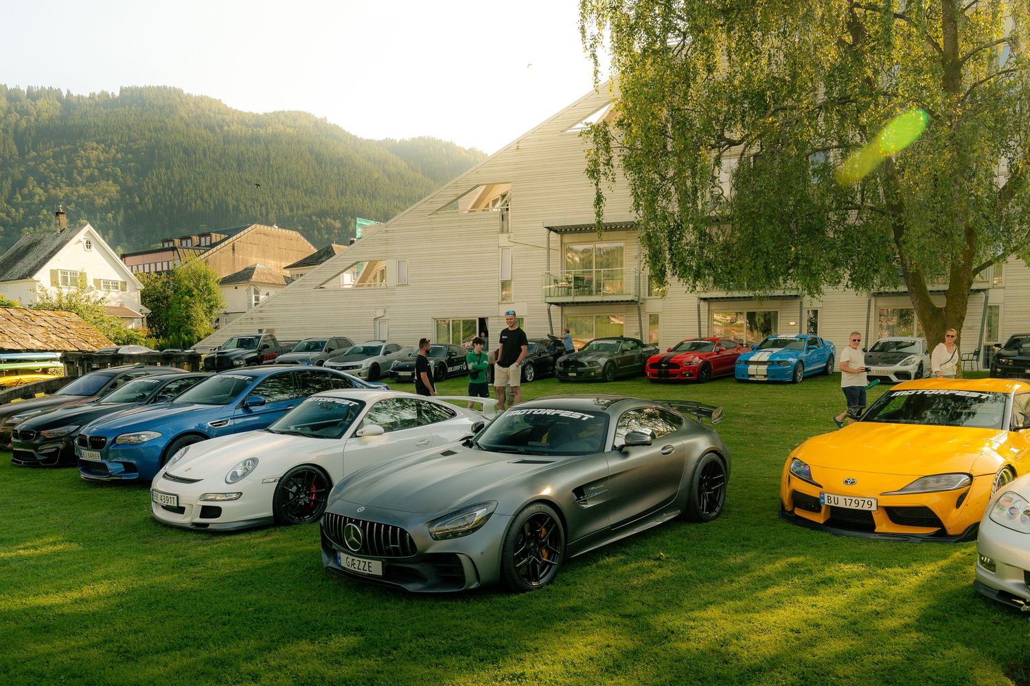 Silver Mercedes-AMG GT R and other sports cars parked on a grassy lawn at an outdoor car show near mountain-side buildings.