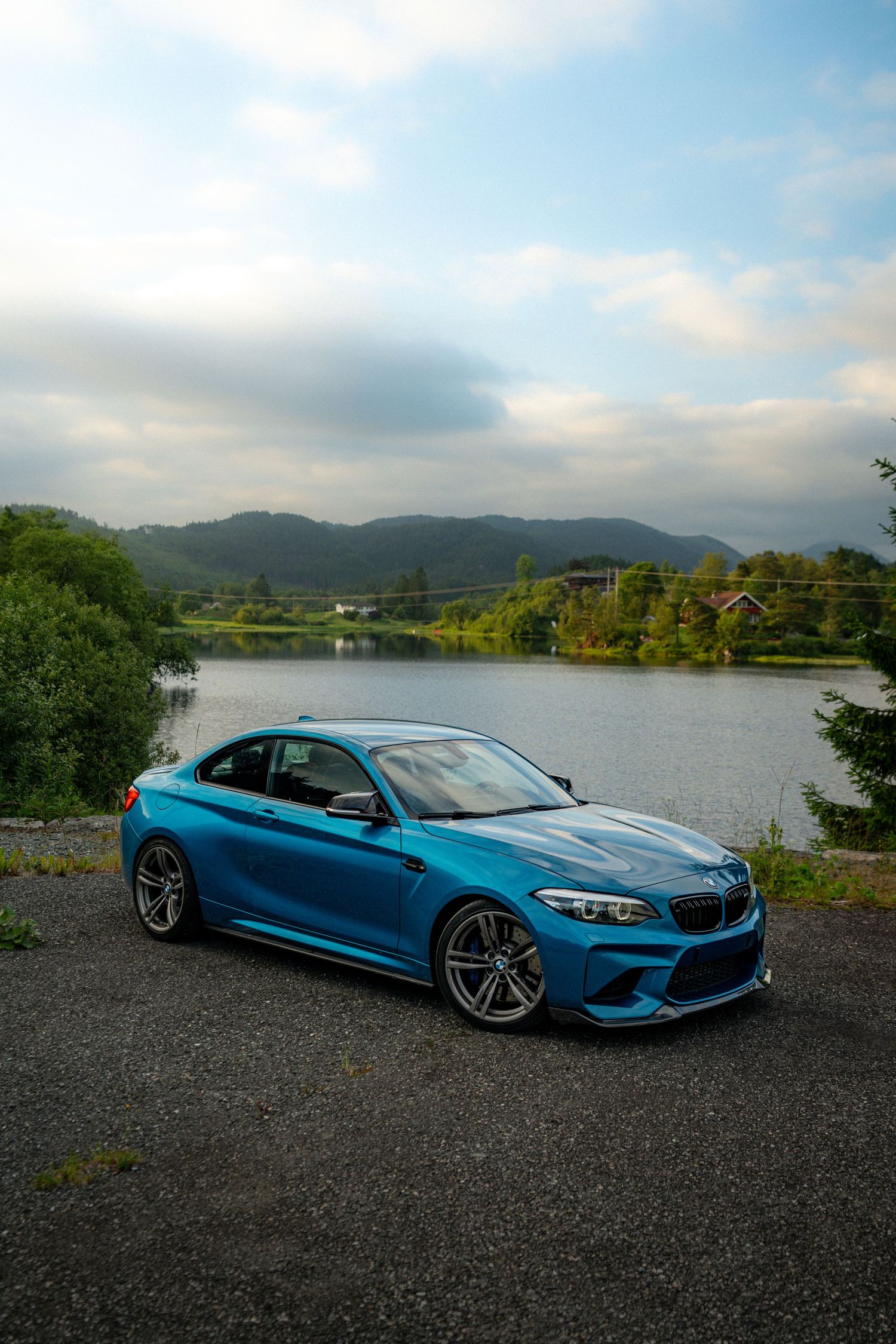 BMW M2 Competition in bright blue finish showcased against serene lakeside landscape.