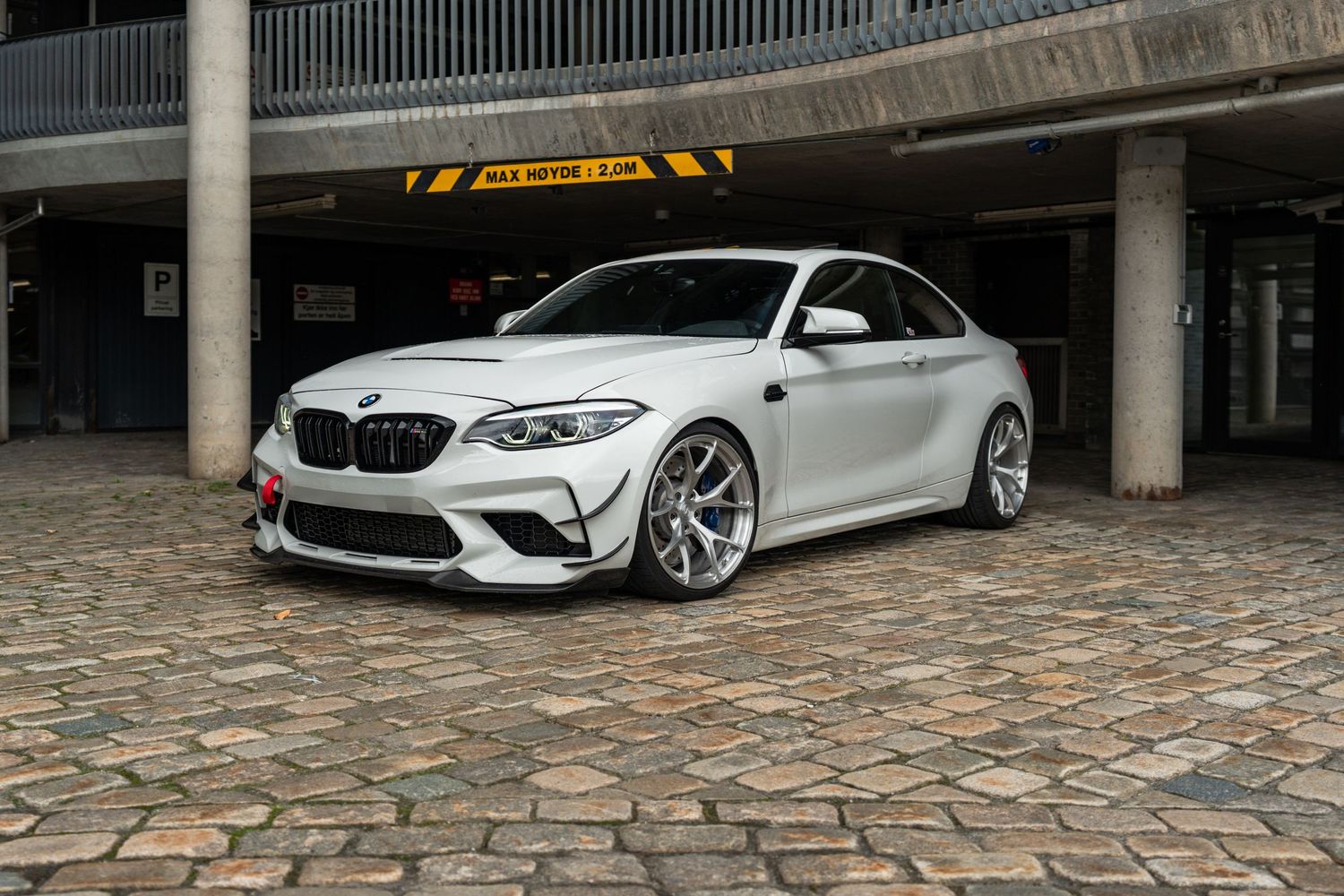 White BMW M2 Competition with custom wheels parked on cobblestone surface.
