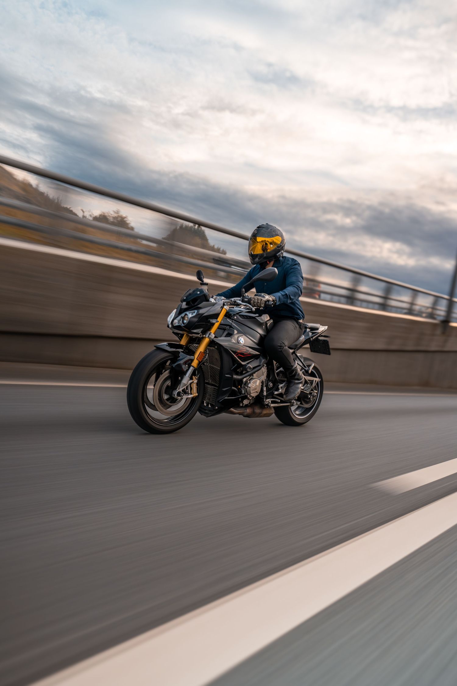 A motorcyclist rides a sporty black and gold bike on a highway during cloudy weather with motion blur effect.