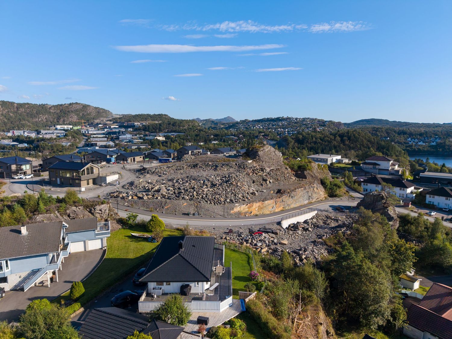 Aerial view of a hillside residential neighborhood with houses, a rocky outcrop, and a coastal town under a clear blue sky.