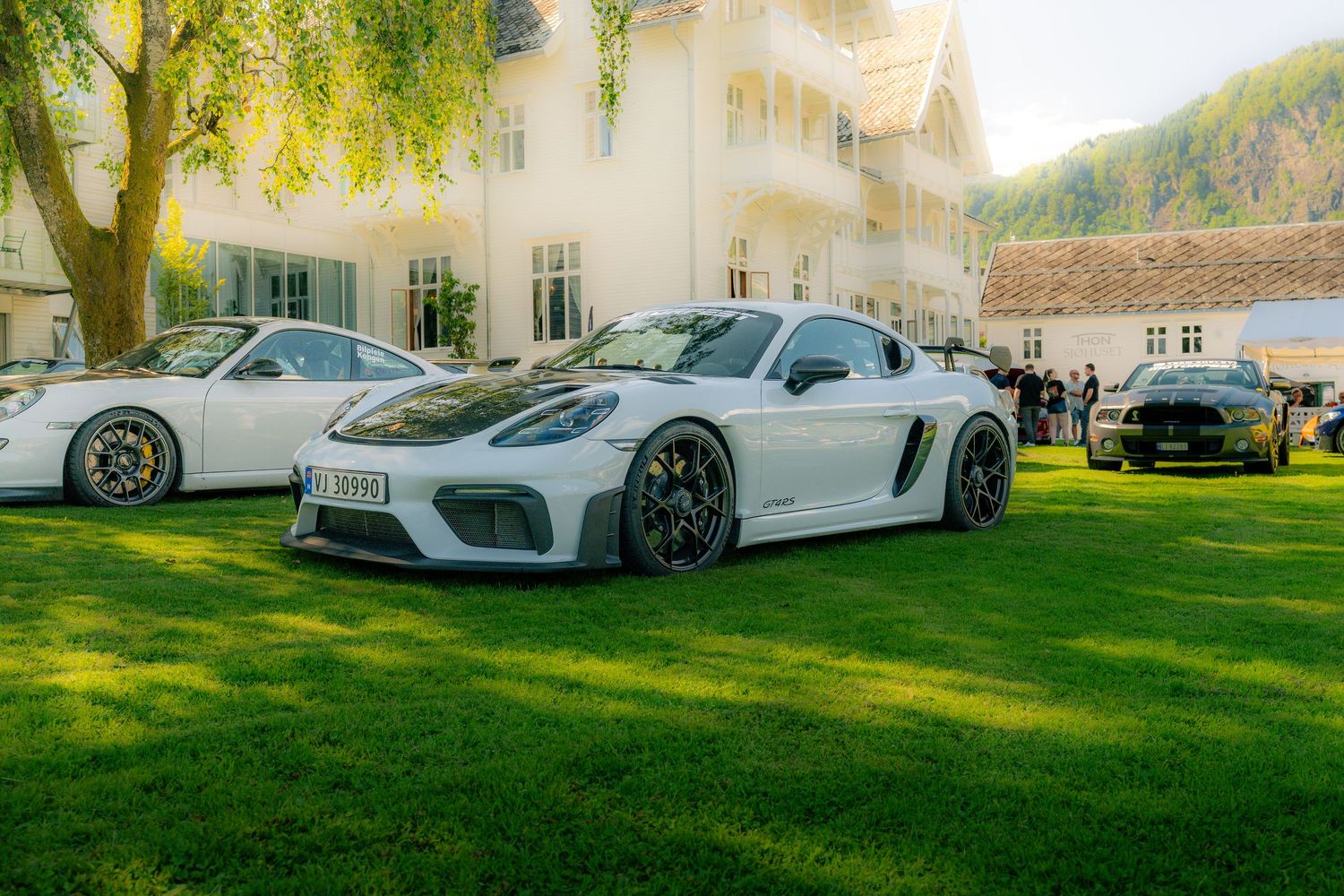 White Porsche 718 Cayman GT4 parked on a sunny lawn at a car meet with houses, trees and mountains in the background.
