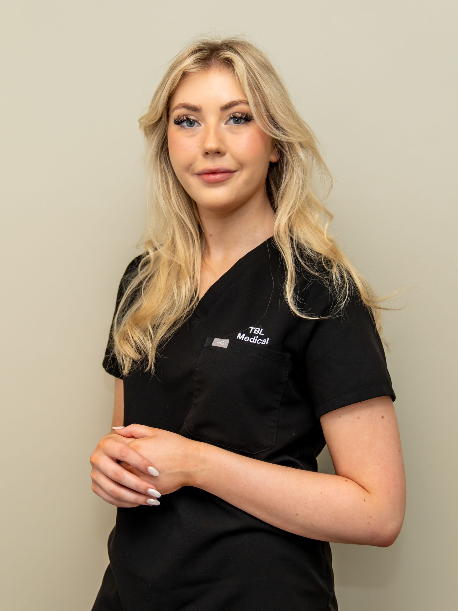 Smiling woman with long wavy blonde hair wearing black TBL Medical scrubs, standing with hands clasped against a neutral beige background.