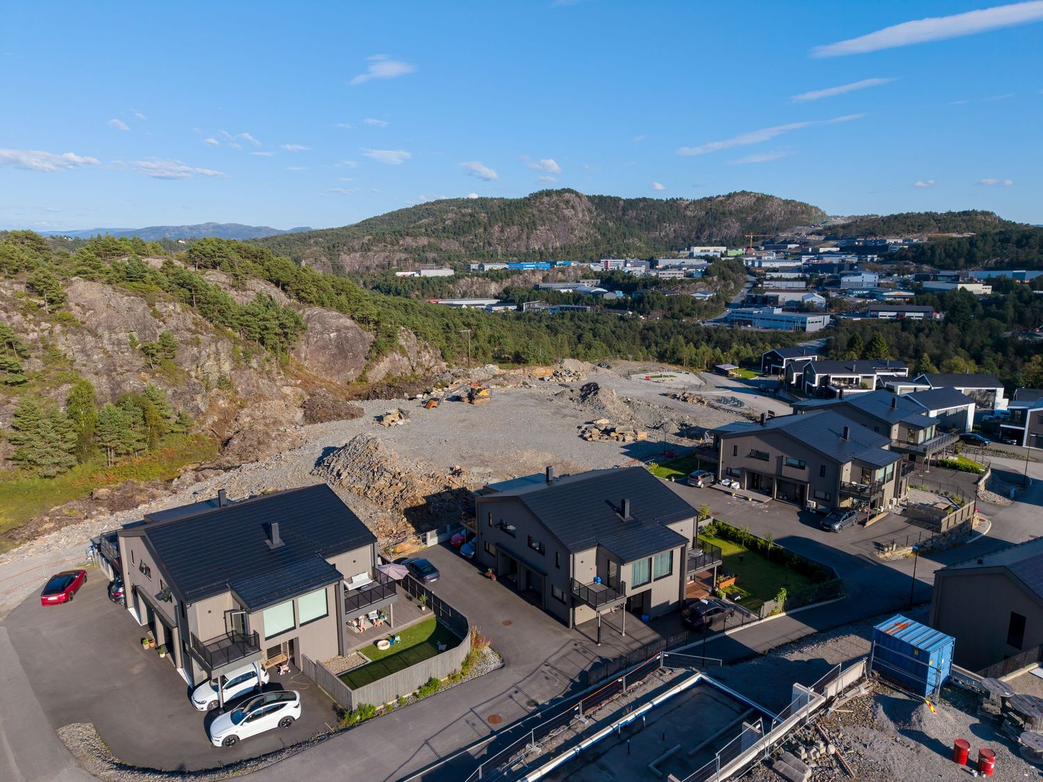 Aerial view of modern dark-roofed houses on a rocky hillside development overlooking a coastal town, forested hills, and a clear blue sky.