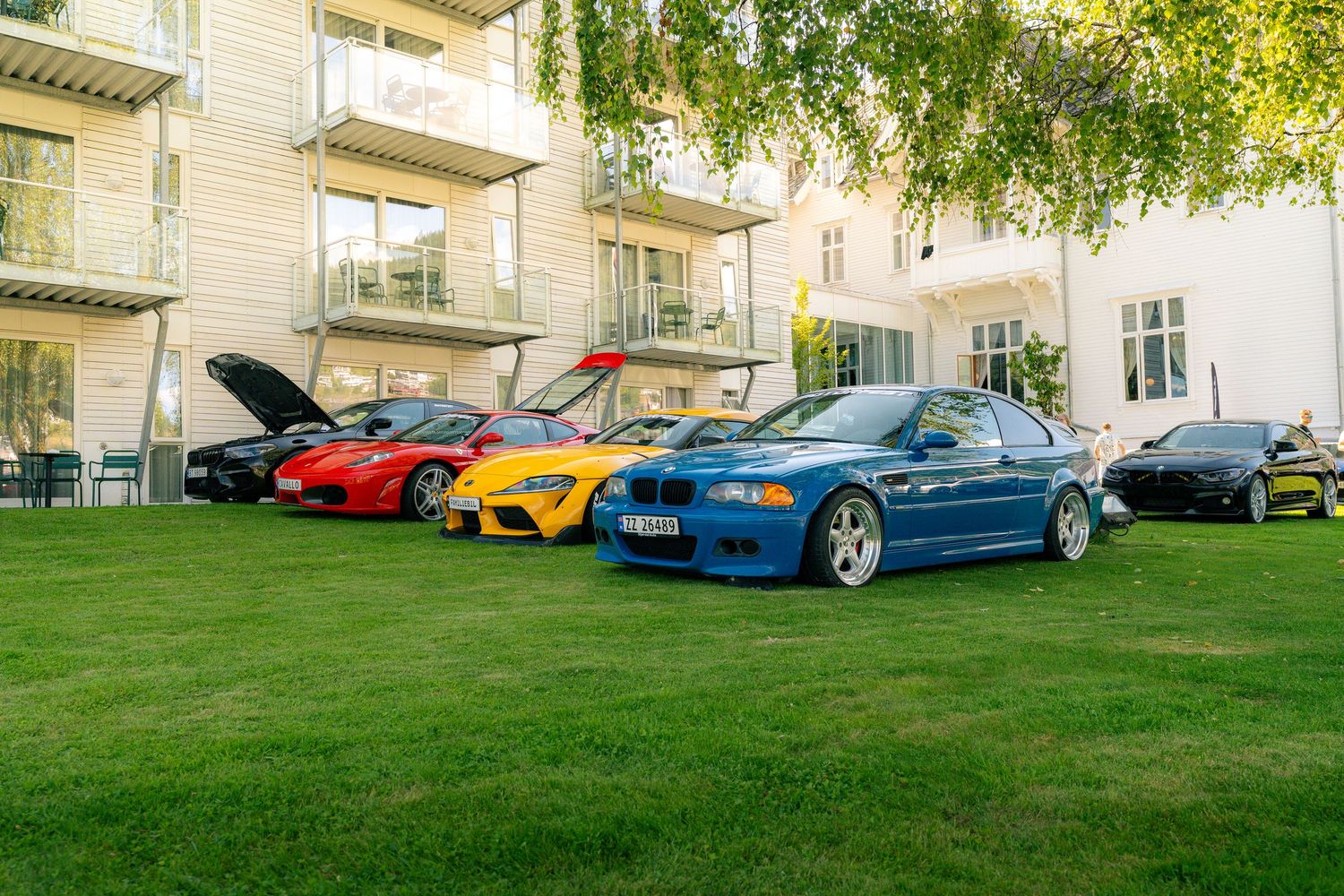 Row of colorful sports cars including a blue BMW coupe, yellow and red models, parked on a green lawn beside a white hotel building.