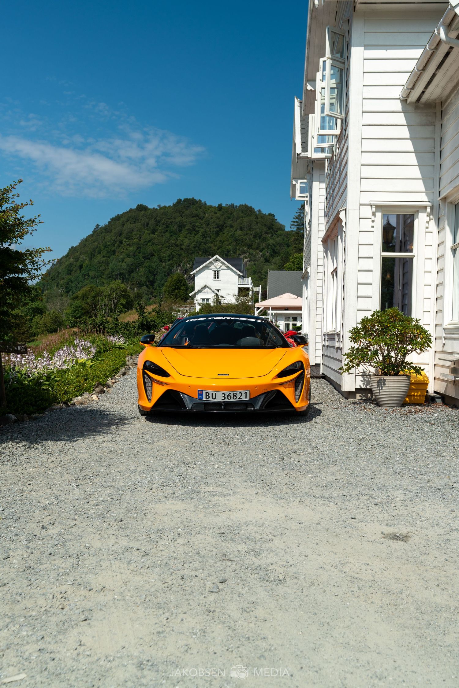 Orange McLaren supercar parked in front of white coastal house with mountains in background.