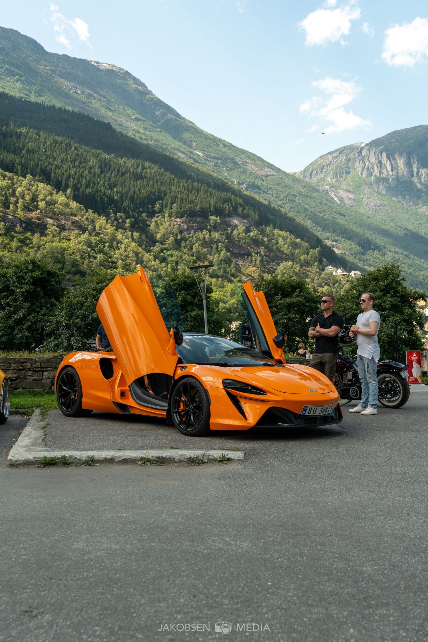 Orange McLaren supercar with dramatic butterfly doors open against a mountainous landscape backdrop.