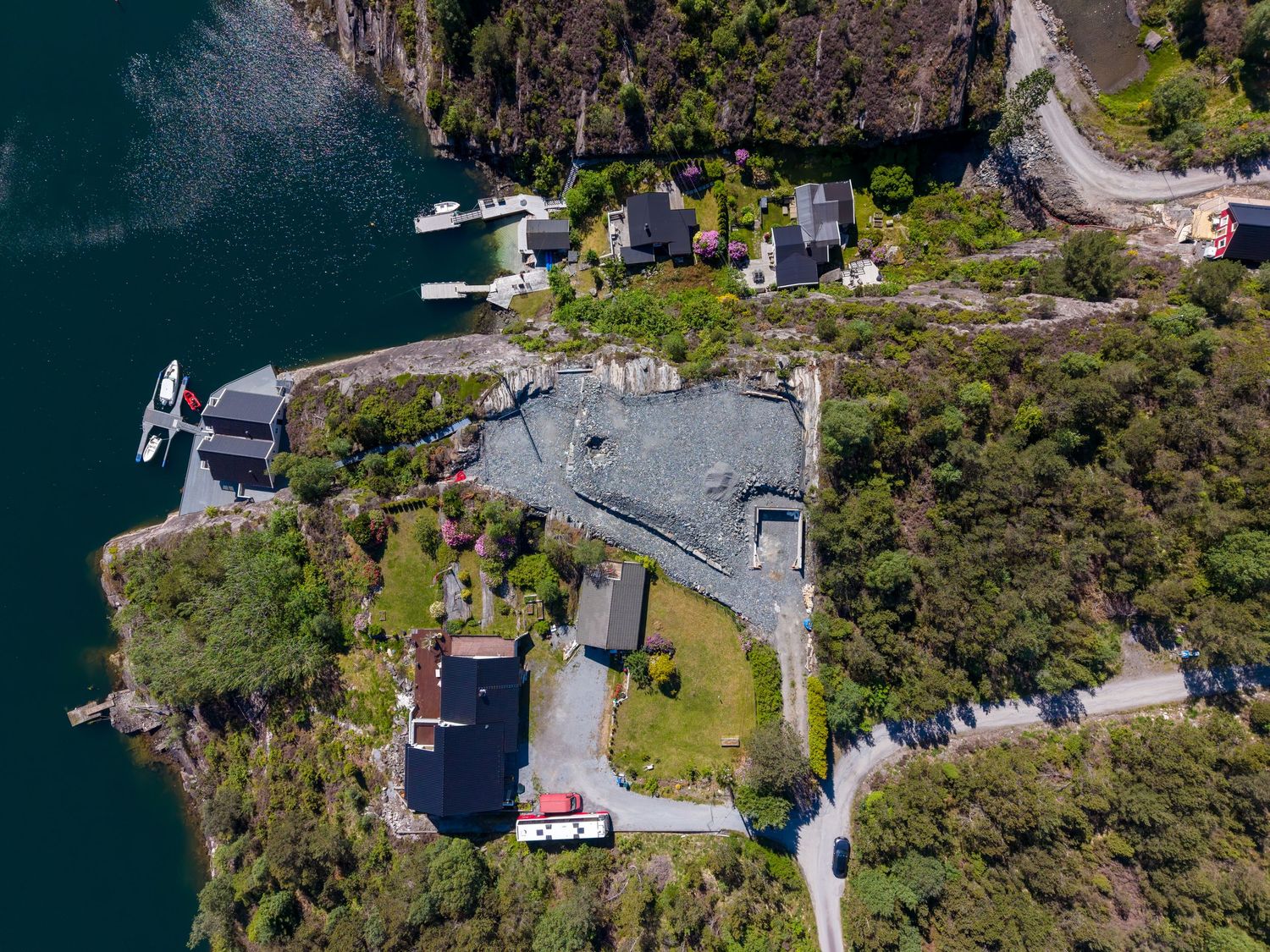 Aerial view of a fjord-side village with houses, a dock with boats, a gray-roofed building, and green forest along the rocky coast.