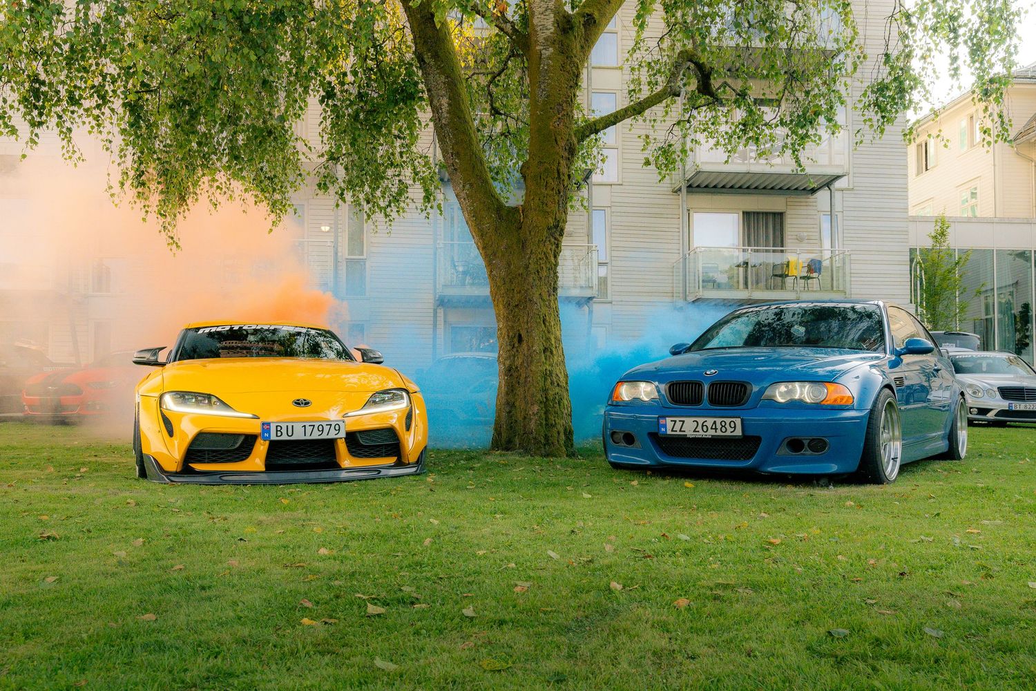 Yellow Toyota Supra and blue BMW M3 parked on grass beneath a willow tree with colorful orange and blue smoke behind.