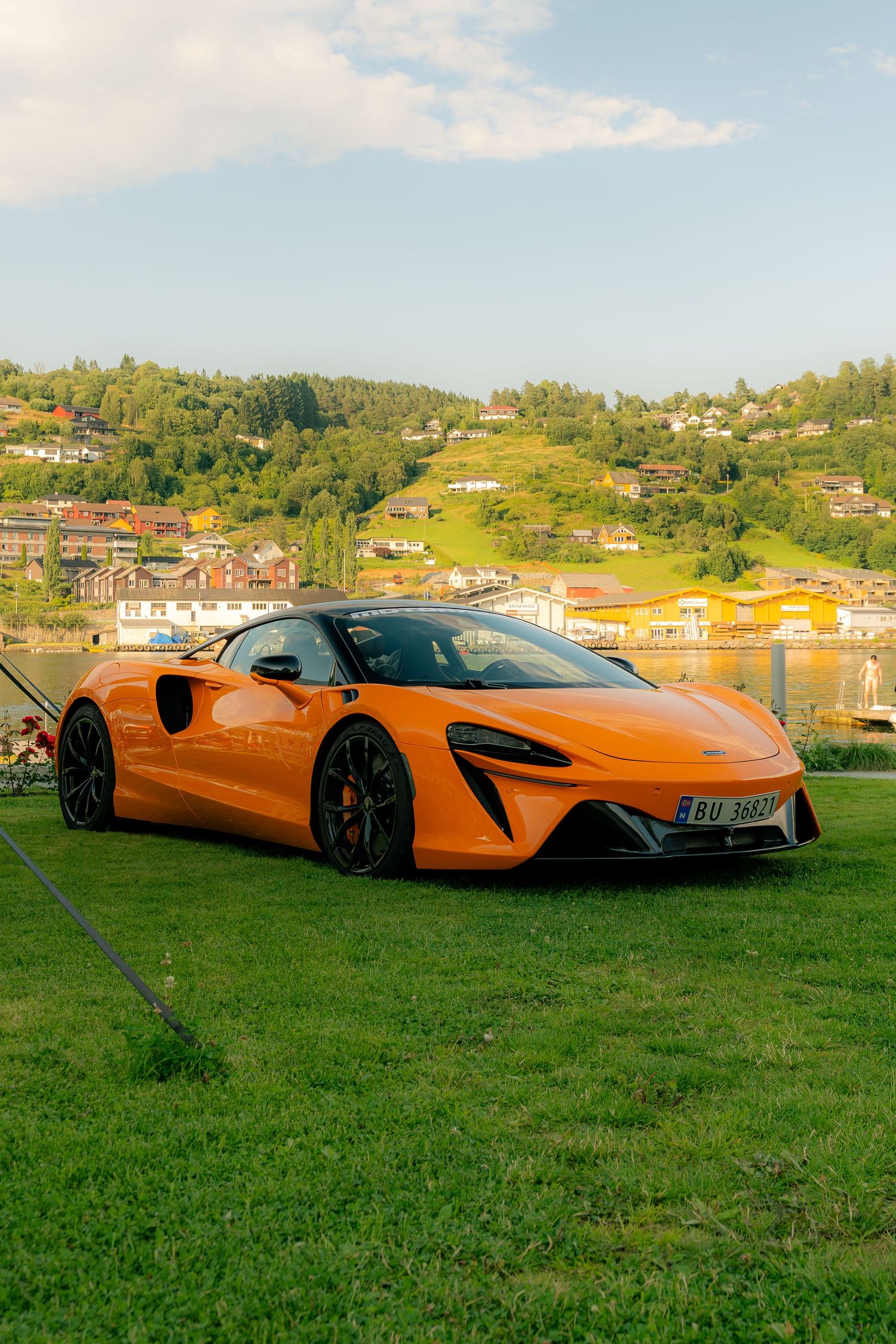Orange McLaren Artura supercar parked on green grass with a scenic lakeside village and forested hills in the background.