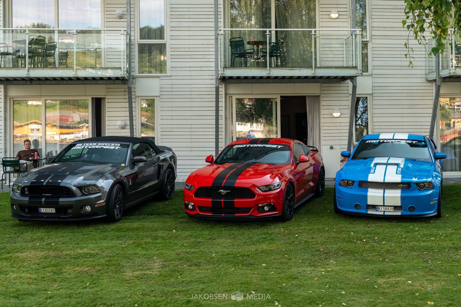 Row of colorful sports cars including red and blue models parked on grass in front of a modern building.