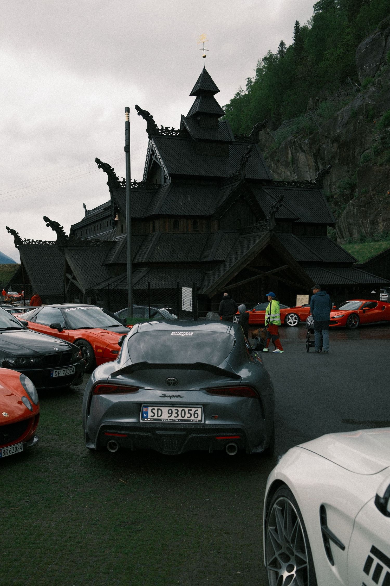 Gray Toyota Supra and other sports cars parked outside a dark wooden stave church with dragon-head gables below a forested mountainside.