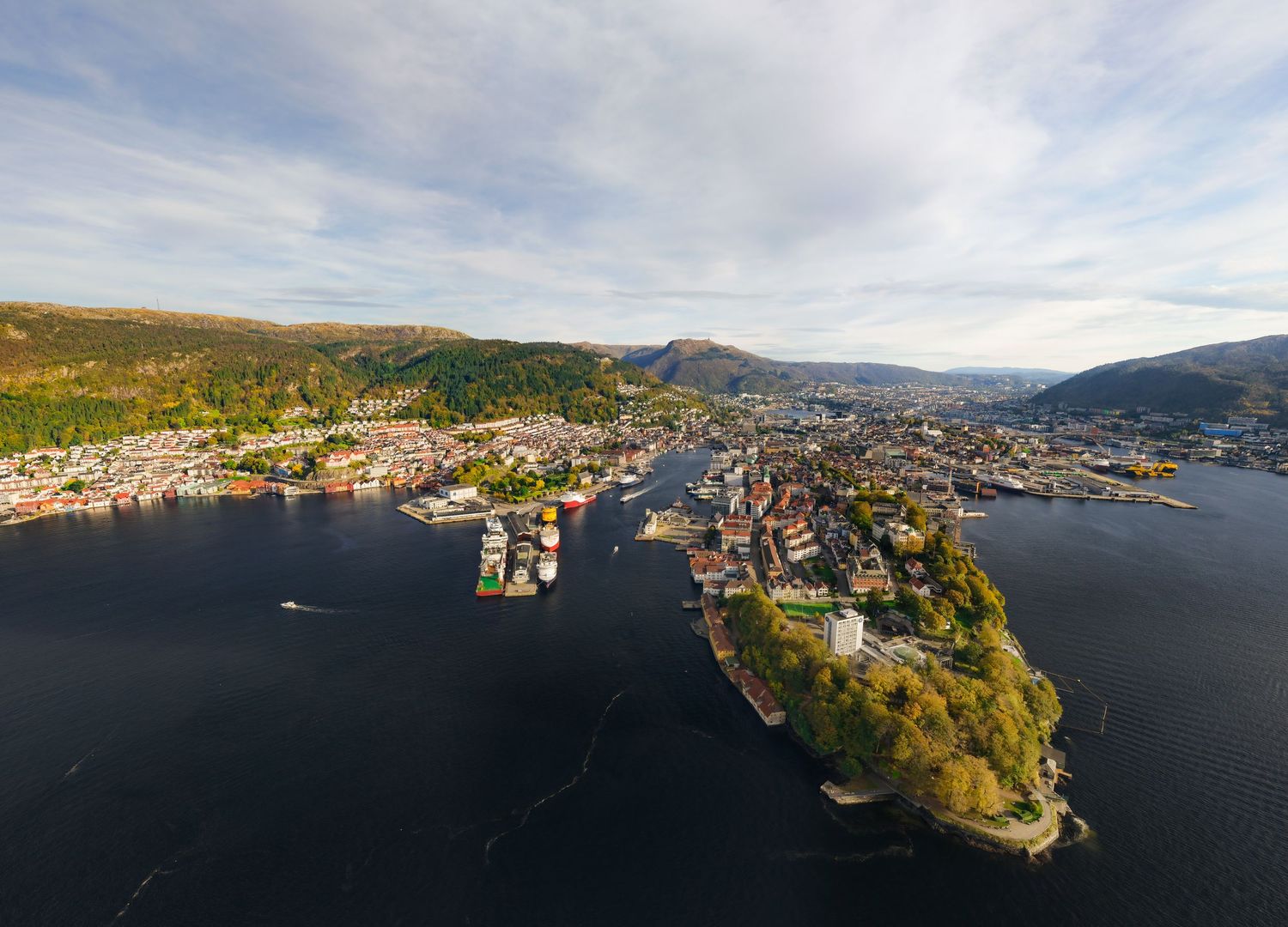 Aerial view of Bergen, Norway, showcasing the city harbor, colorful buildings, and lush green hills under a partly cloudy sky.