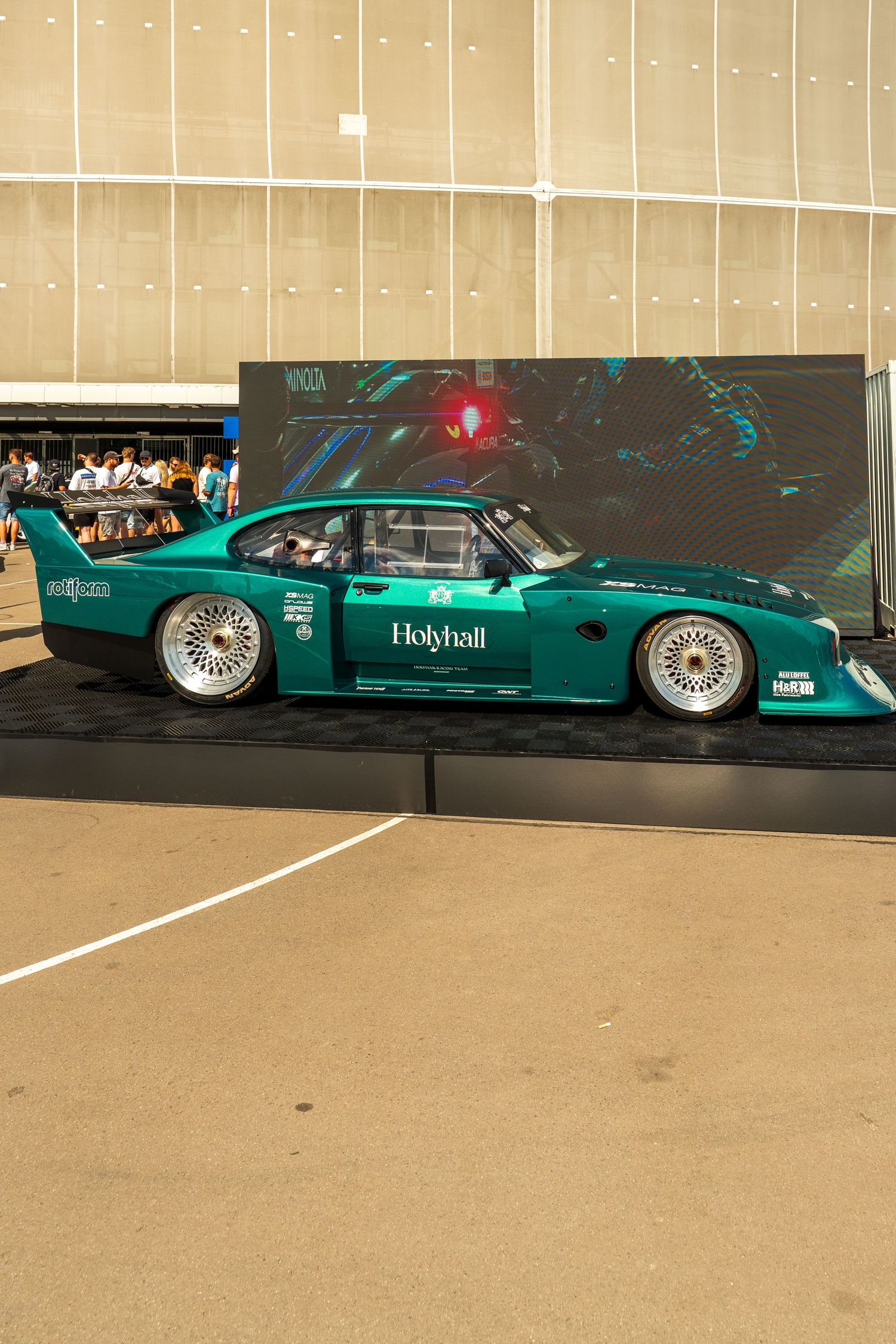 Modified teal racing car with custom body kit and aerodynamic features parked in front of dark wall.