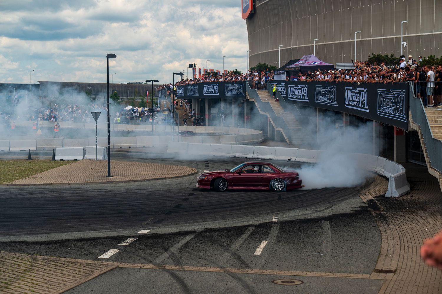 Red sports car performing a burnout with smoke while crowd watches from above.