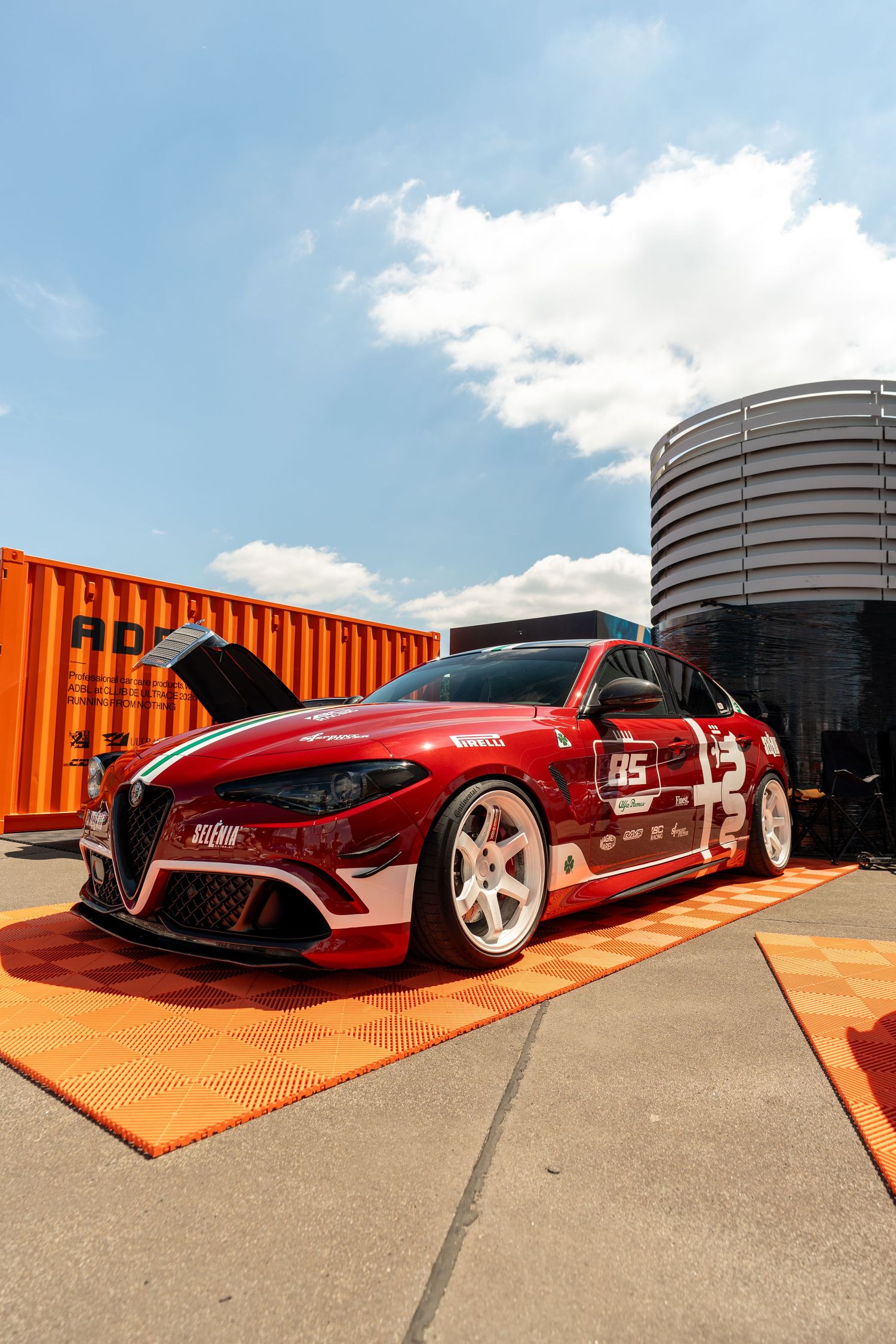 Custom red sports car with racing stripes parked on orange platform outdoors.